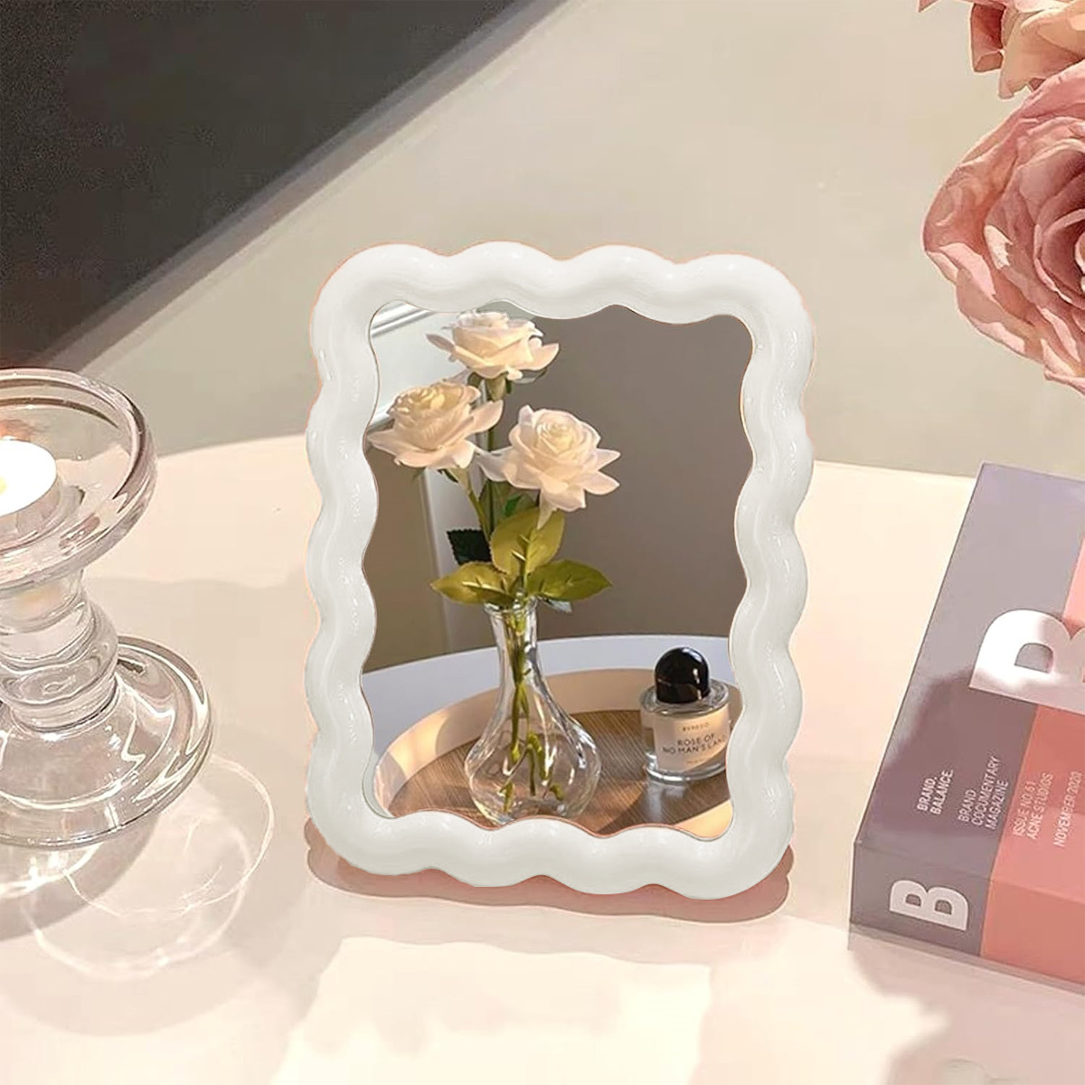 Ein wellenförmiger weißer Spiegel mit einer Vase Rosen, einer Kerze und einem Buch. Heller Hintergrund.