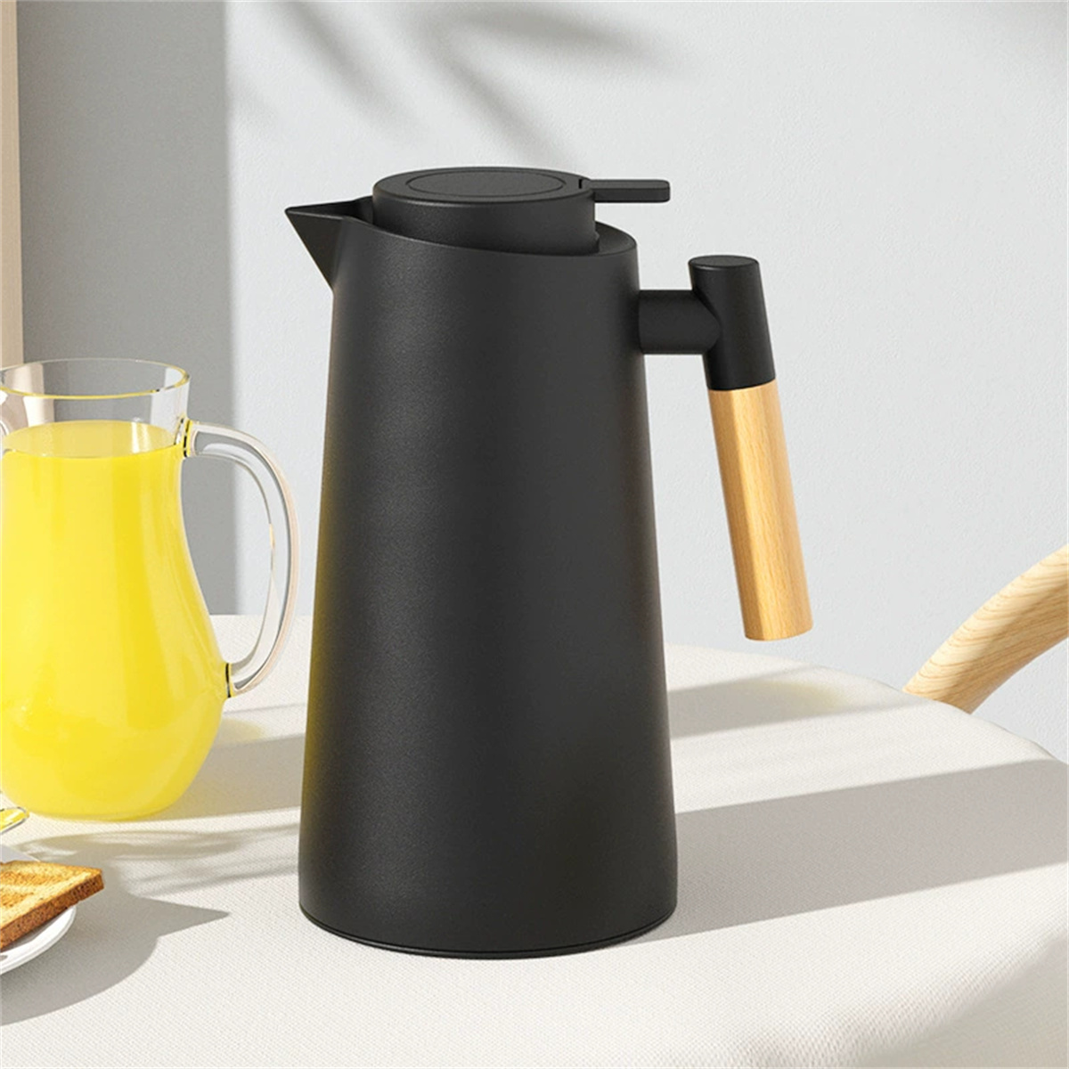 Eine schwarze Thermoskanne mit Holzgriff, auf weißer Oberfläche, neben einem Glas und einem Teller mit Essen.