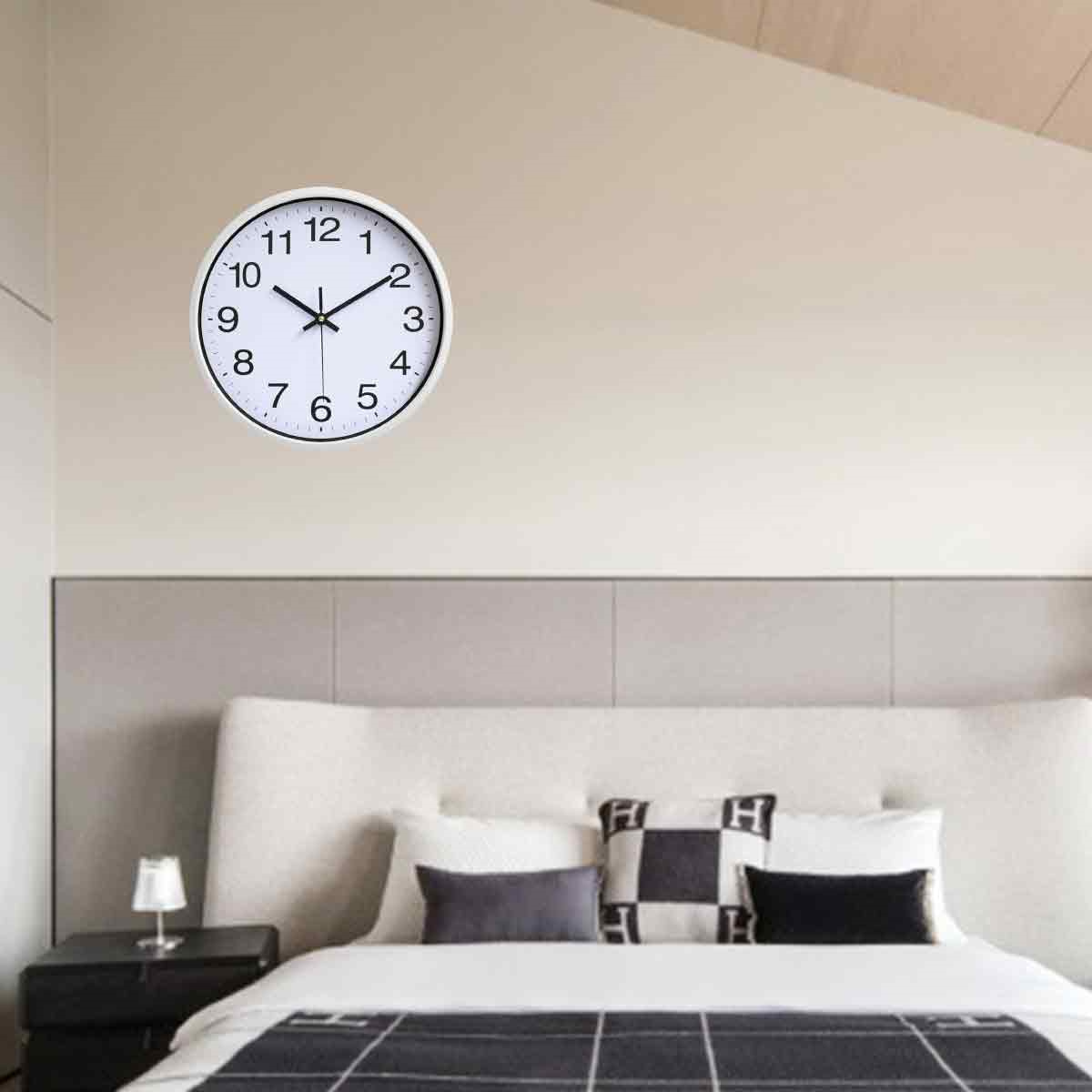 Eine runde, weiße Wanduhr an einer weißen Wand. Im Hintergrund befinden sich ein Bett und Kissen.