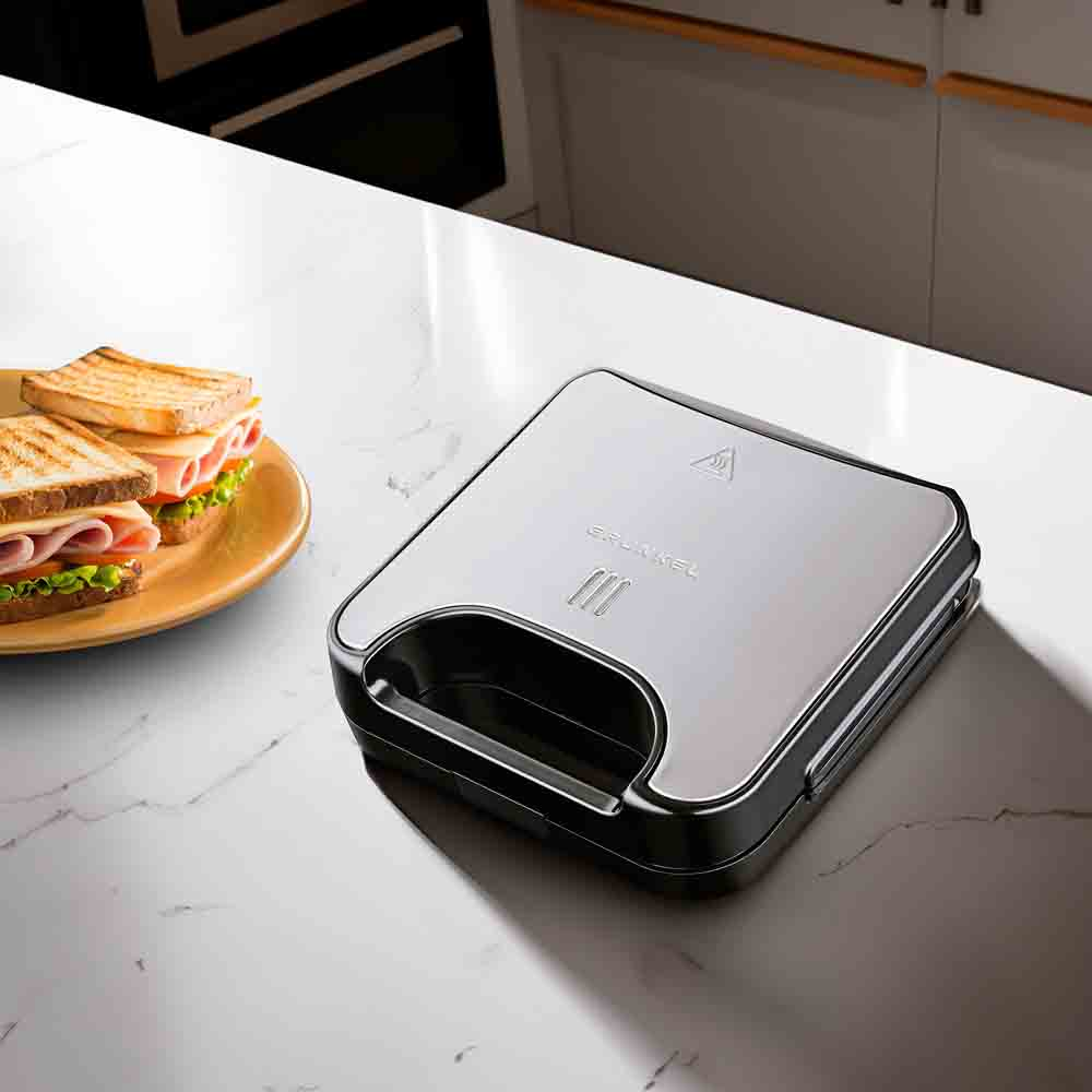 Una macchina per sandwich argentata e nera e un piatto con panini su un piano di lavoro bianco.
