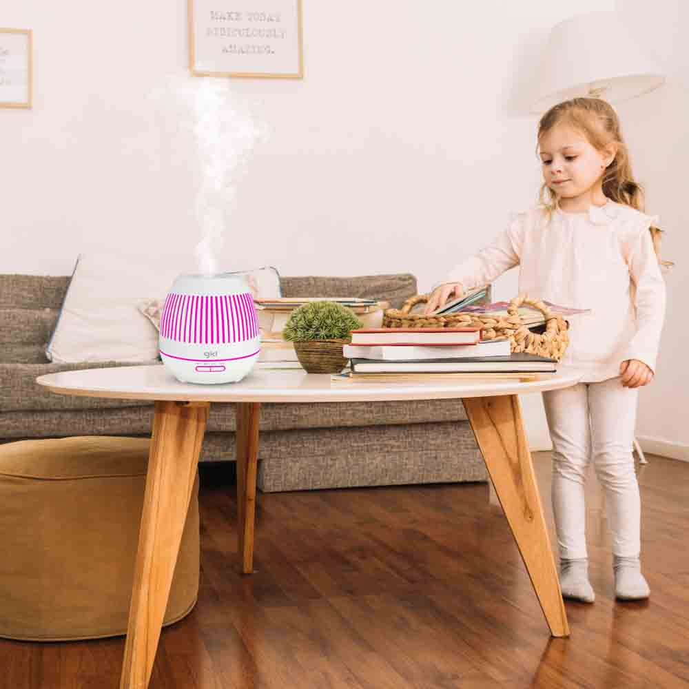 Una bambina è in piedi vicino a un tavolo con un diffusore che emette vapore. Libri e una pianta sono sul tavolo.
