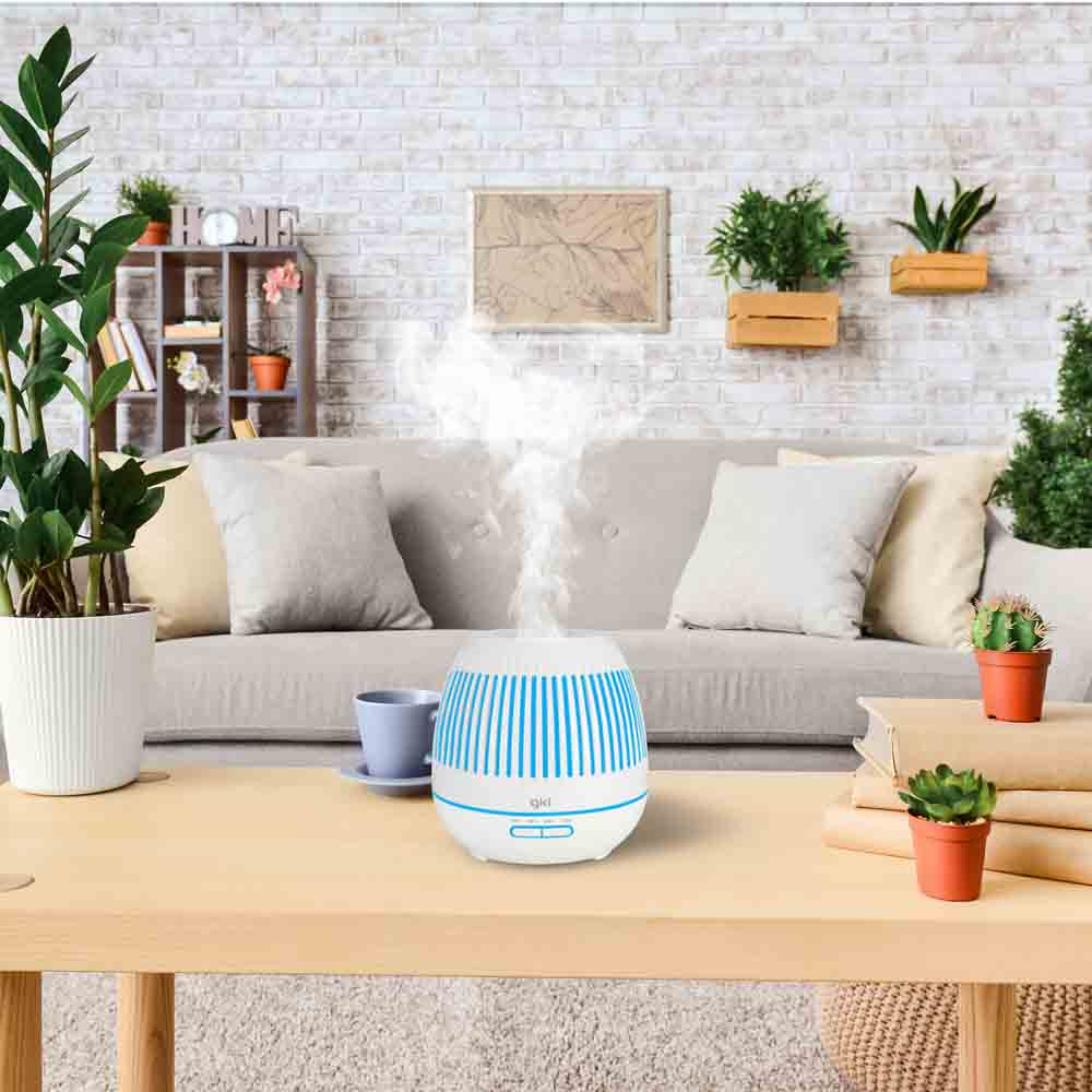 Diffusore di aromi su un tavolo di legno, vapore che sale. Piante e divano sullo sfondo. Blu e bianco.