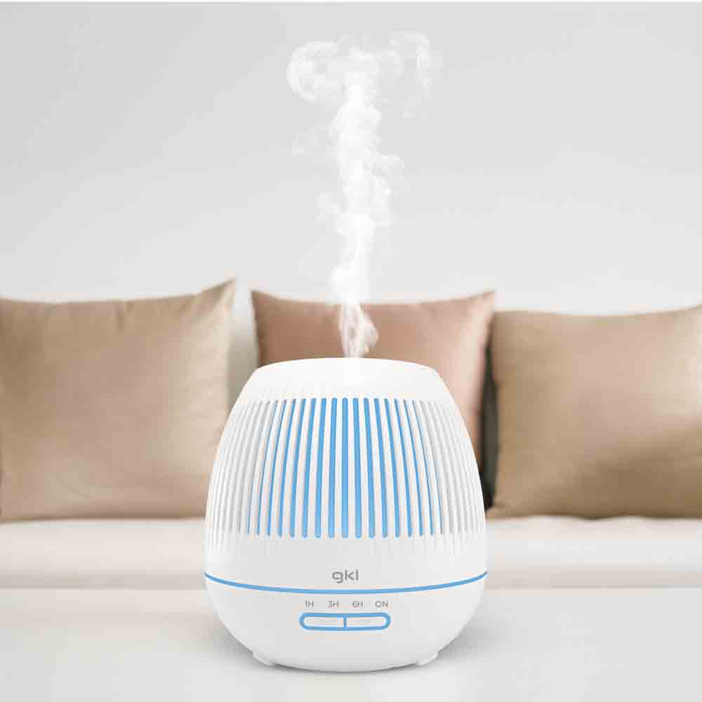 Diffusore di aromi bianco e blu. Il vapore sale, appoggiato su un tavolo. Cuscini beige sullo sfondo.