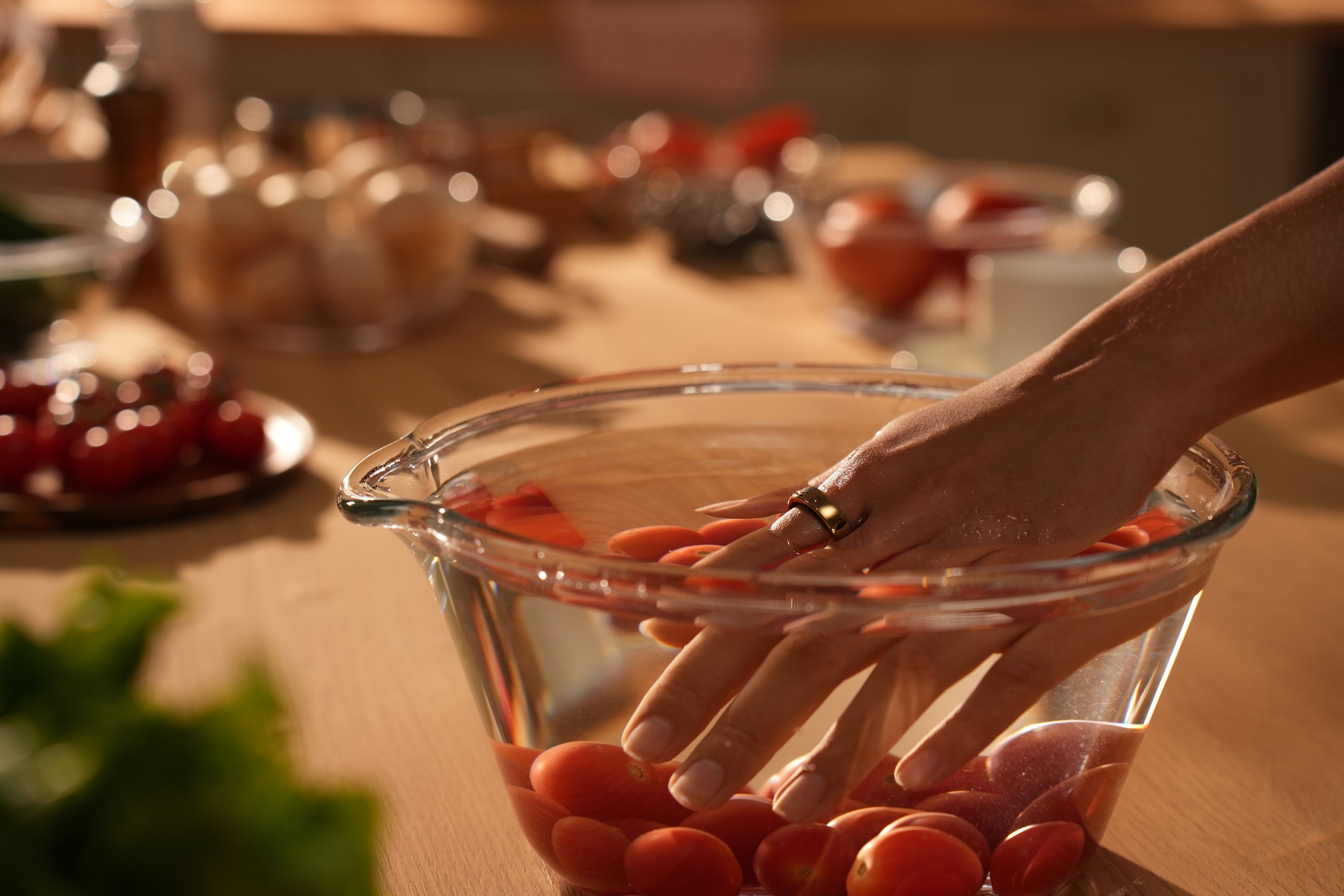 Eine Hand in einer Schüssel mit Wasser und Tomaten, neben einem Holztisch mit Tomaten und Grün.