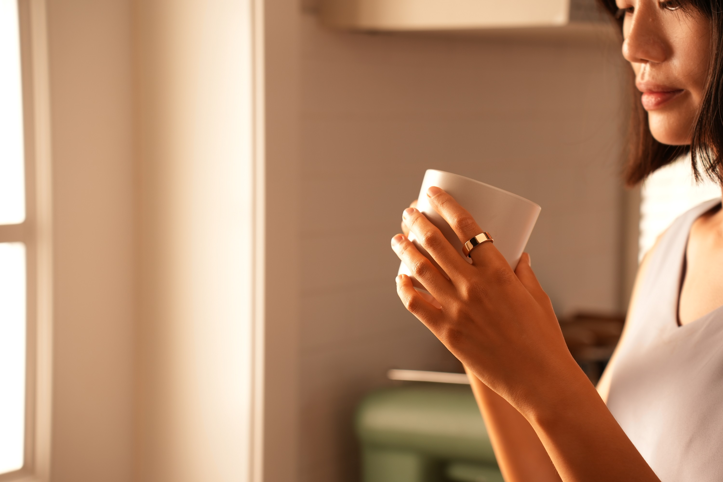 Frau hält eine weiße Tasse in den Händen. Sie trägt einen Ring und ein helles Oberteil. Hintergrund ist unscharf.