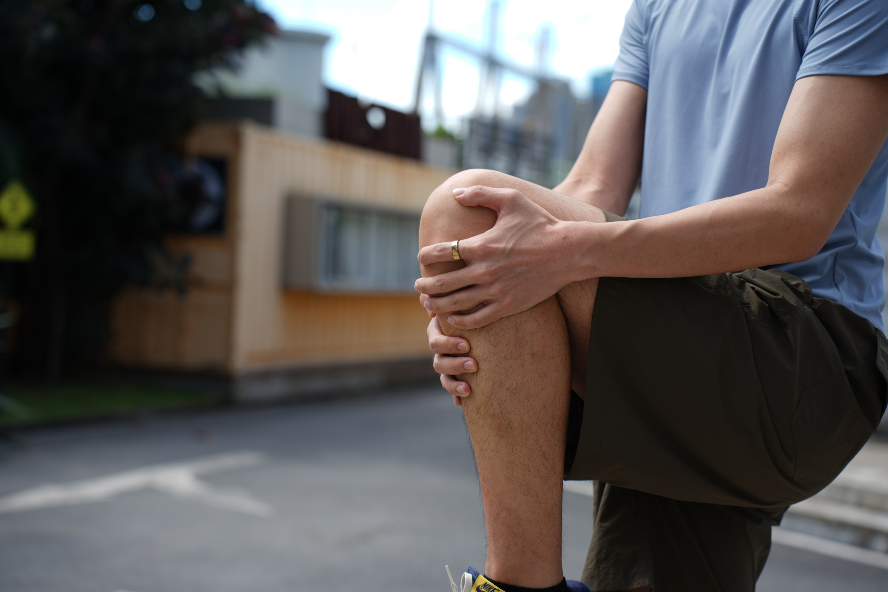 Ein Mann in Shorts hält sein Knie, in einer Umgebung im Freien. Der Hintergrund ist unscharf.