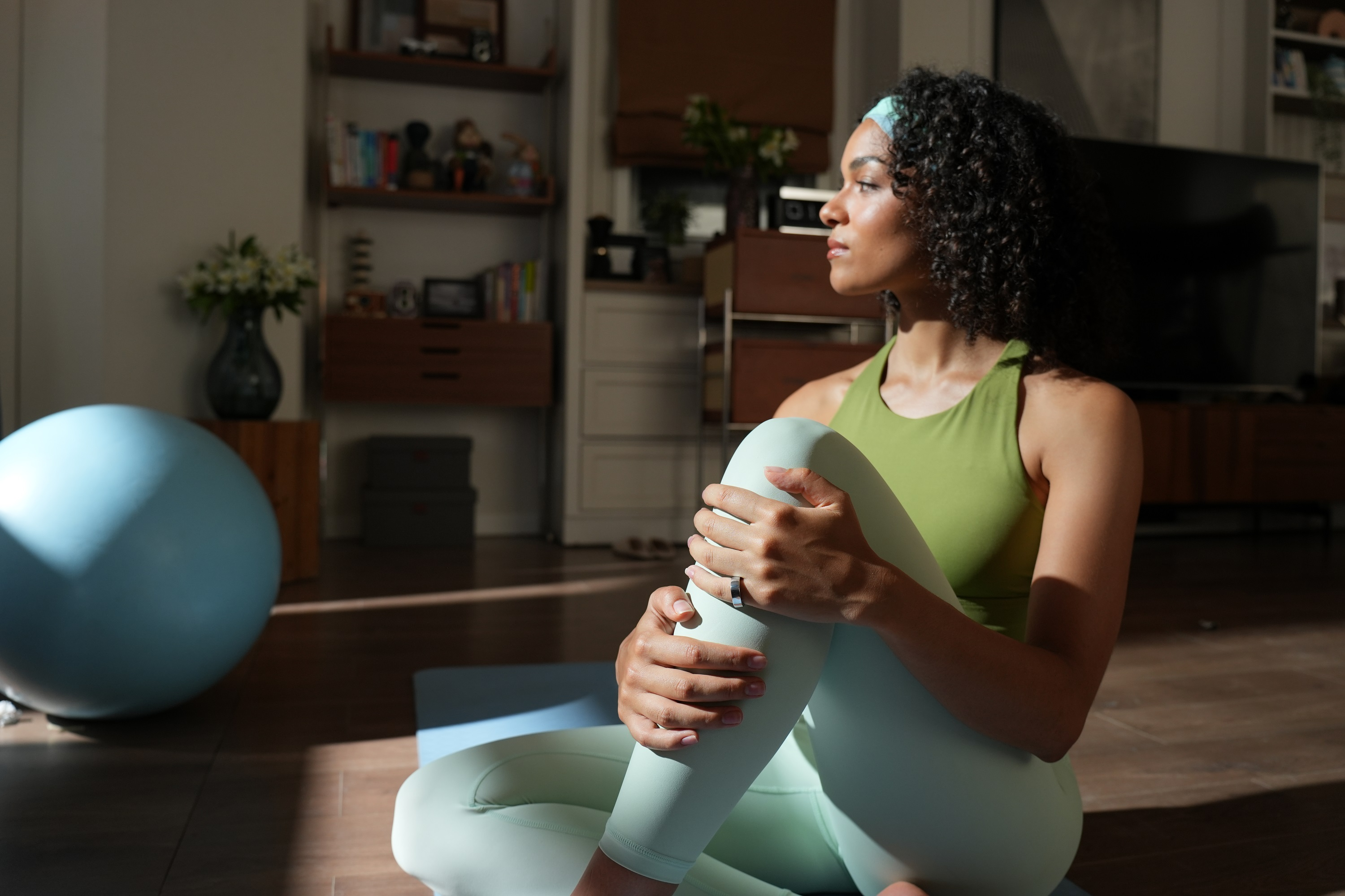Frau in grünem Top und Leggings, auf Matte sitzend, dehnend, mit blauem Gymnastikball.