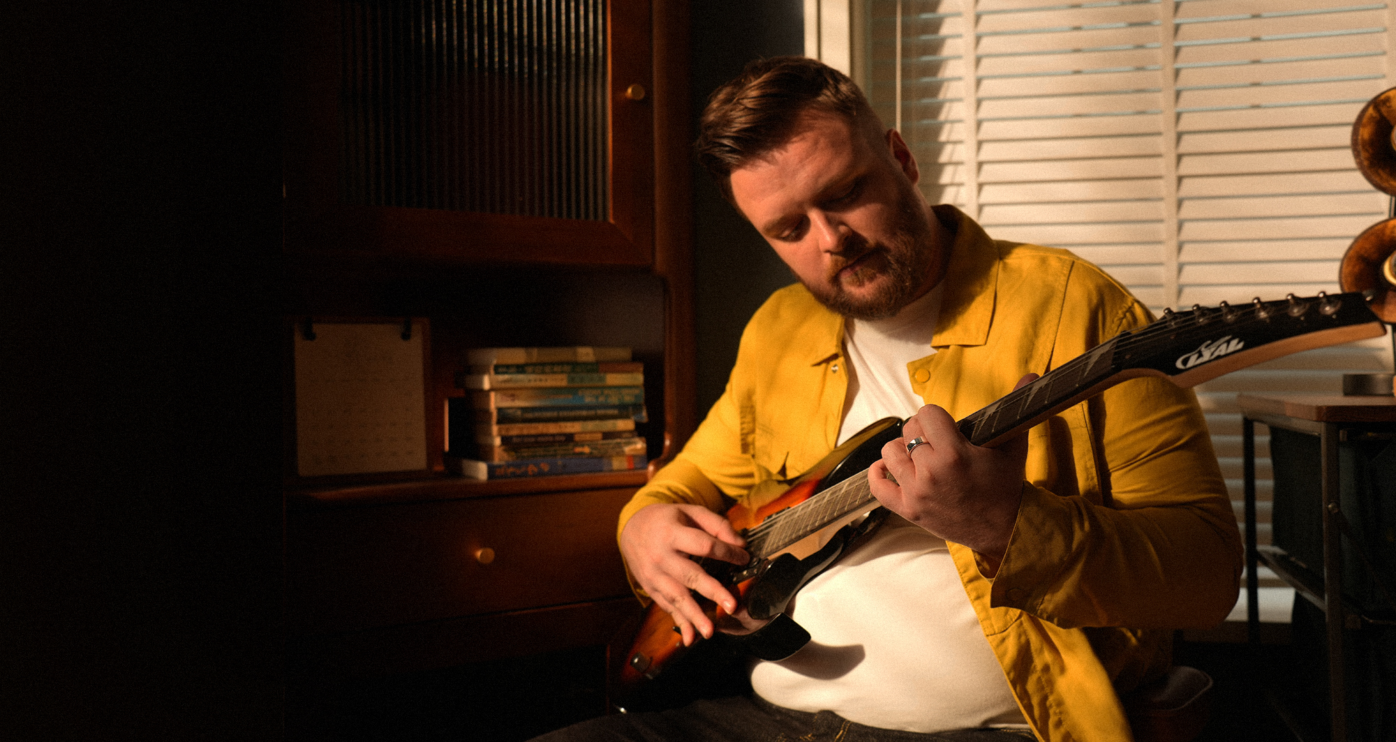 Ein Mann in gelbem Hemd spielt E-Gitarre in einem Raum mit Büchern.
