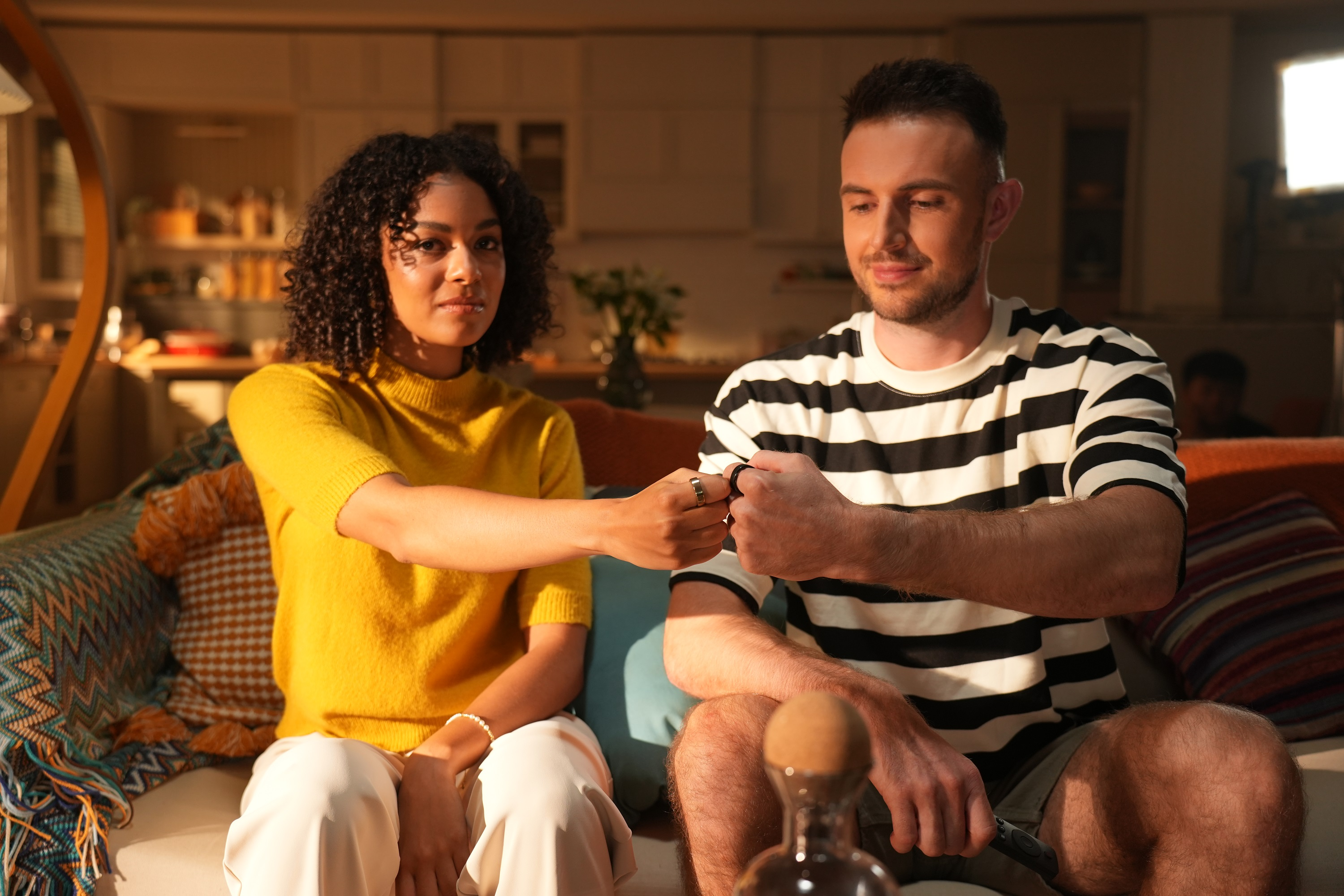 Ein Mann und eine Frau sitzen auf einem Sofa, stoßen Fäuste an, im Hintergrund eine Flasche.