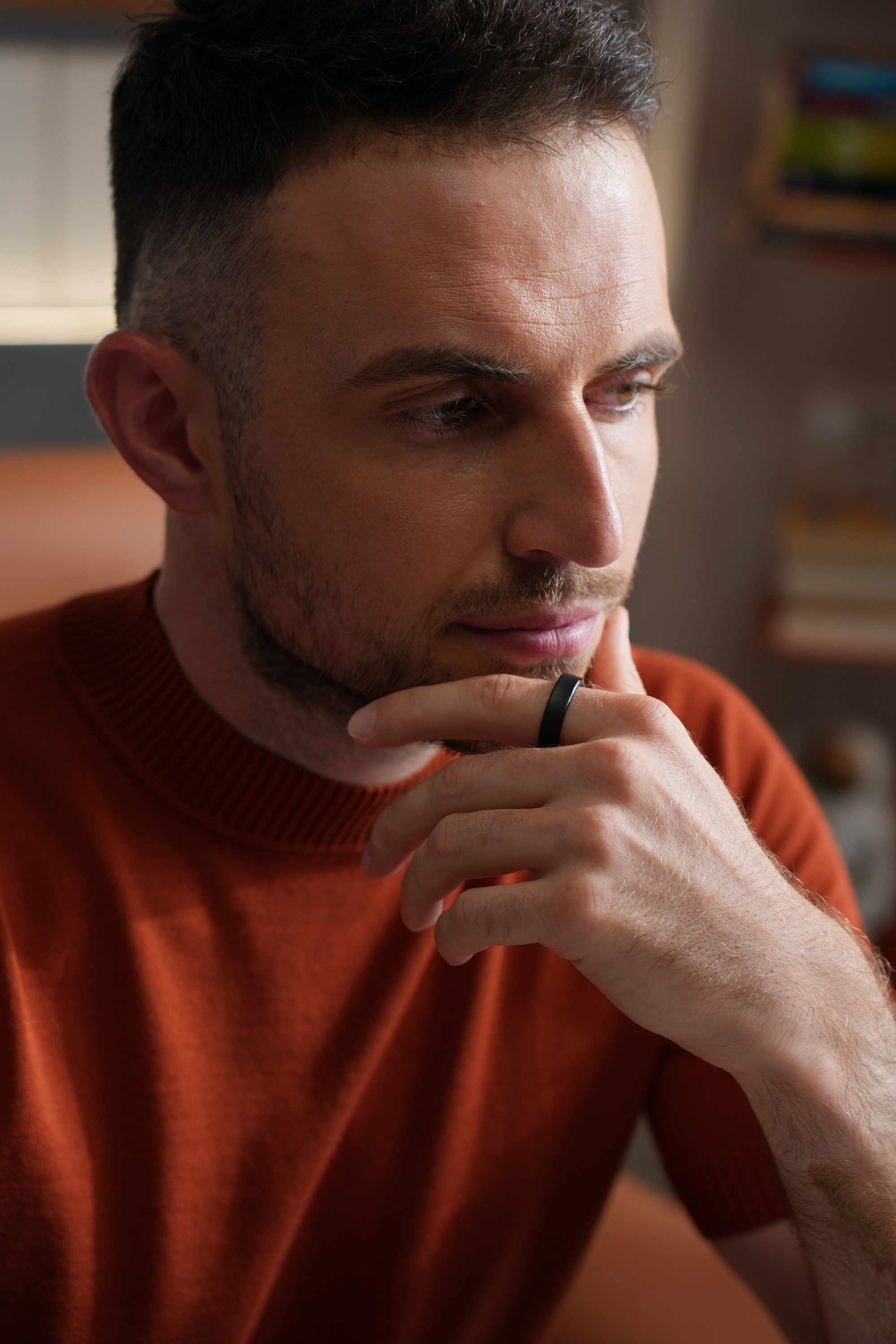 Mann im orangefarbenen Pullover, nachdenklich, berührt sein Gesicht, schwarzer Ring am Finger.