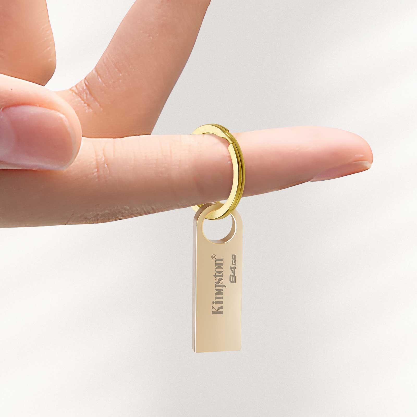 Dłoń trzyma złotą pamięć USB na pierścieniu. Ma logo Kingston i 64 GB. Tło jest białe.