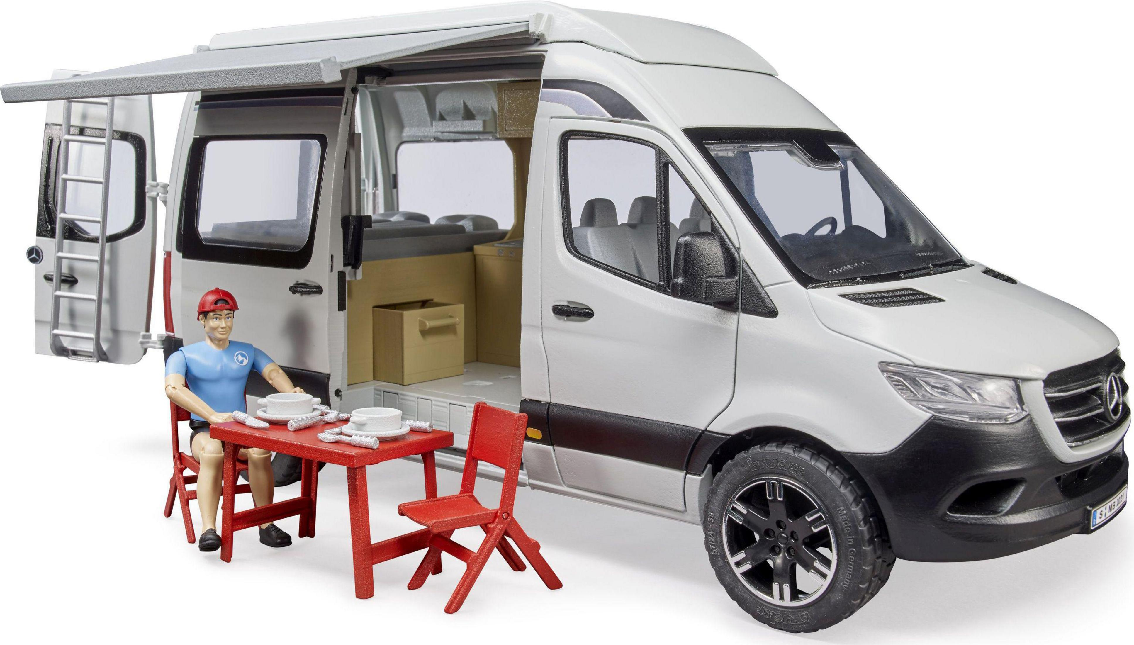 Biały kamper z otwartymi drzwiami, czerwony stół/krzesła, siedzący mężczyzna. Słoneczny dzień.