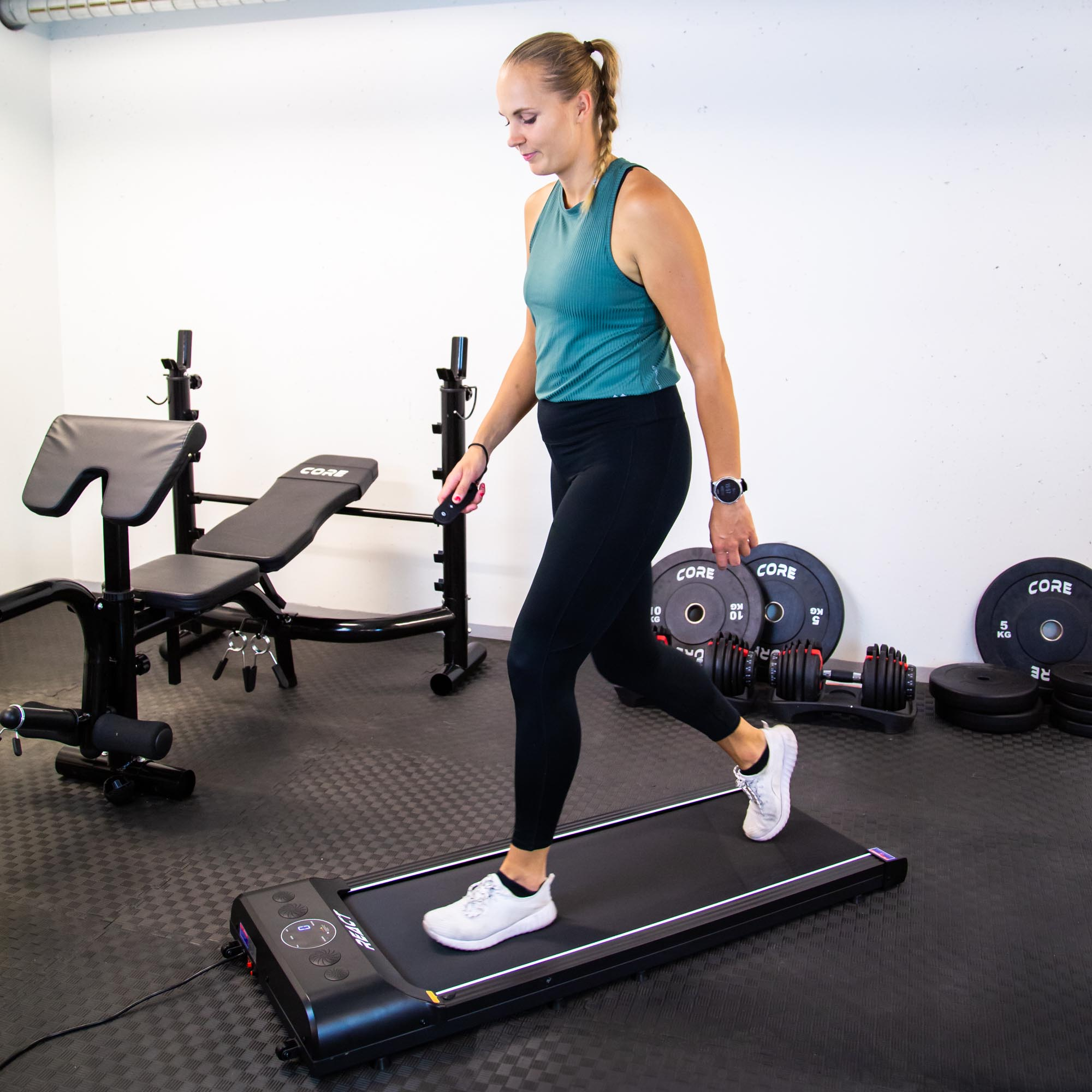 Kobieta idzie po czarnej bieżni na siłowni. Obciążniki i ławka w tle.