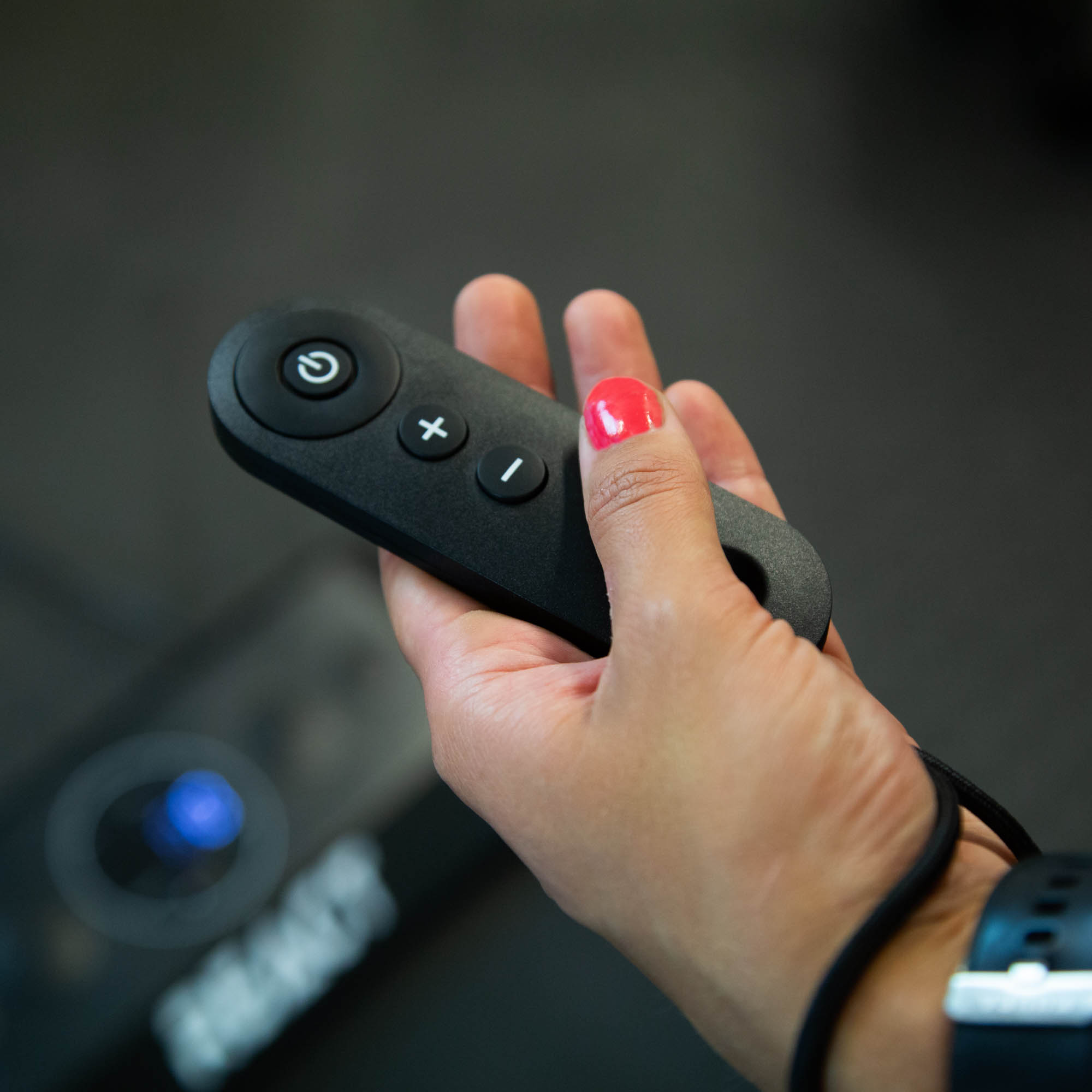 Ręka trzyma czarny pilot z przyciskami zasilania i głośności.