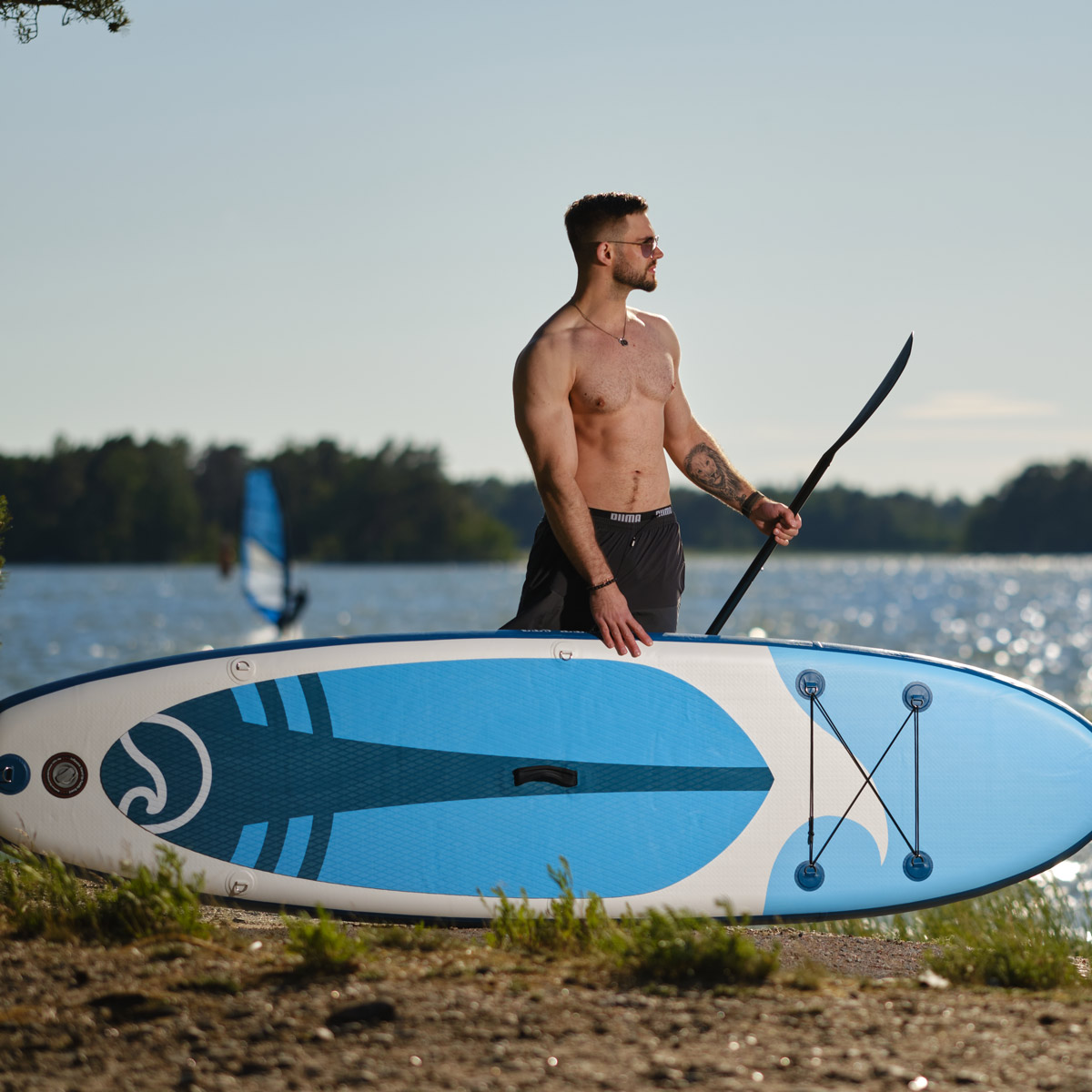 Mężczyzna bez koszuli z deską do wiosłowania na plaży, jezioro w tle. Widoczny windsurfer.
