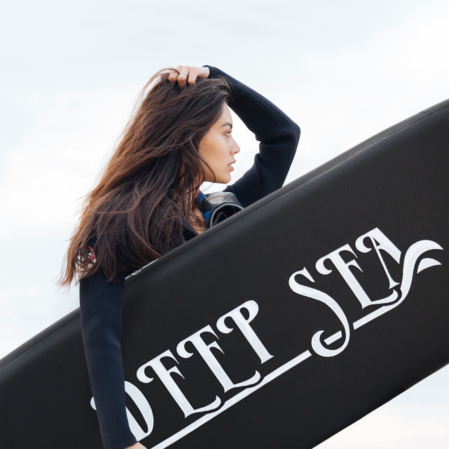 Kobieta z długimi, ciemnymi włosami trzymająca czarną deskę surfingową z napisem 'SOFT SEA'. Pochmurne niebo.