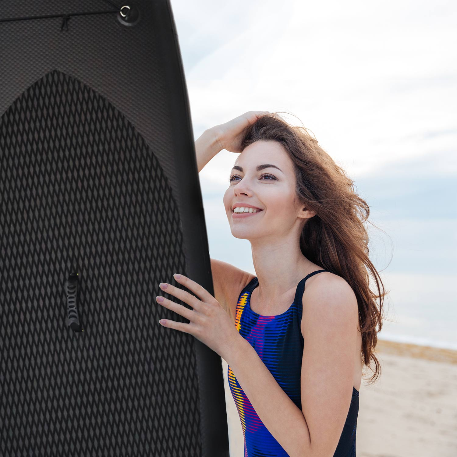 Kobieta uśmiecha się obok deski surfingowej na plaży. Niebo jest czyste i widać morze.