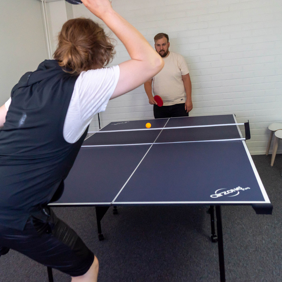 Zwei Männer spielen Tischtennis. Einer schwingt einen Schläger, der andere schaut zu. Ein gelber Ball liegt auf dem Tisch.