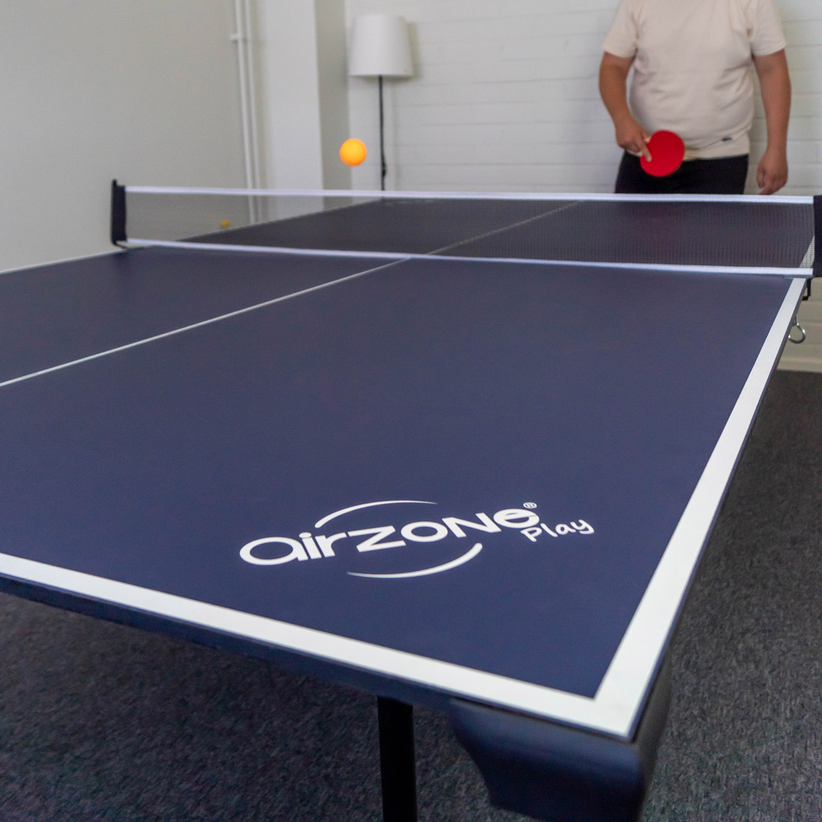 Ein marineblauer Tischtennistisch mit schwarzem Netz. Ein Mann spielt Tischtennis. Ein orangefarbener Ball ist in der Luft.