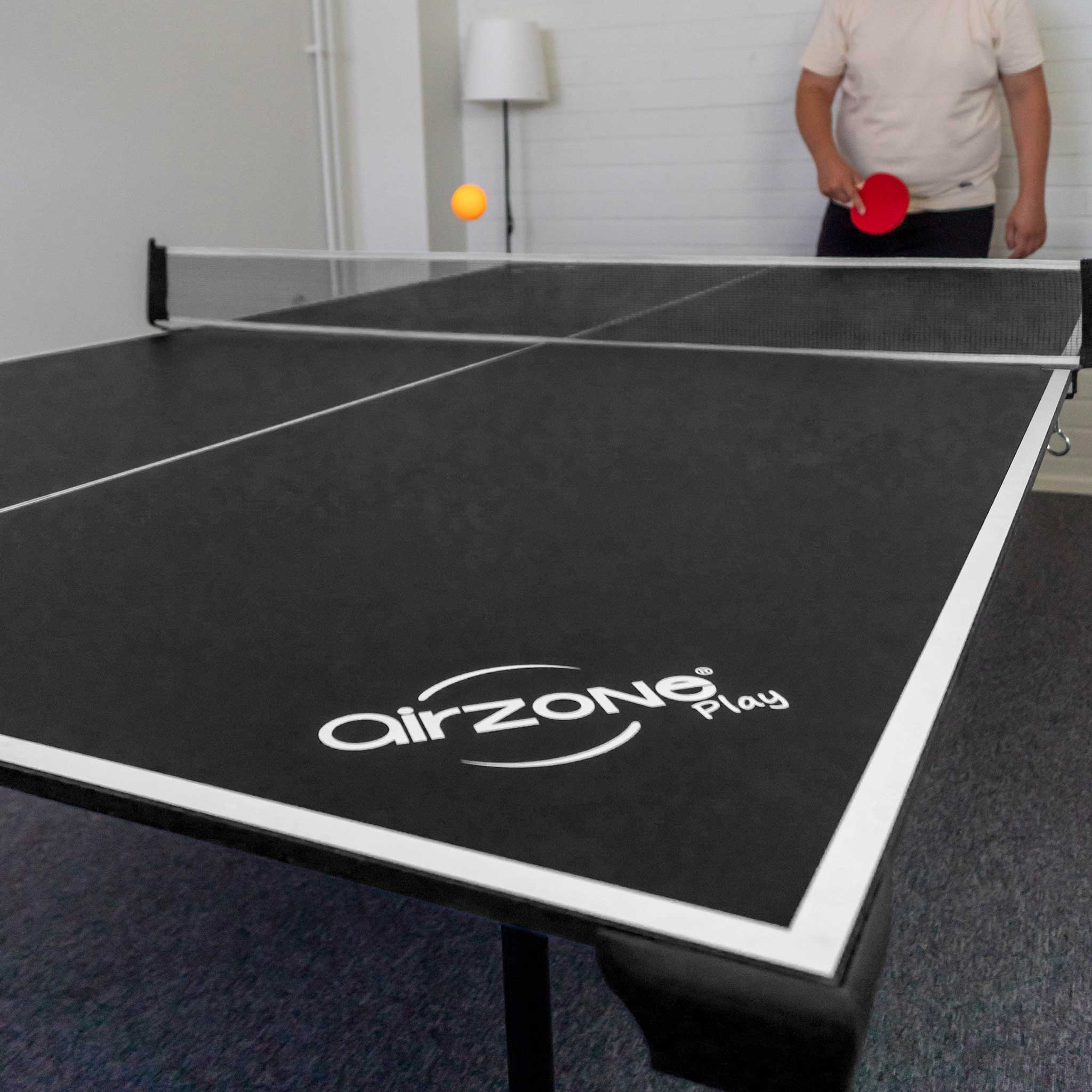 Tischtennisplatte mit Ball in der Luft und einer Person mit Schläger. Schwarzer Tisch mit weißen Linien und Logo.