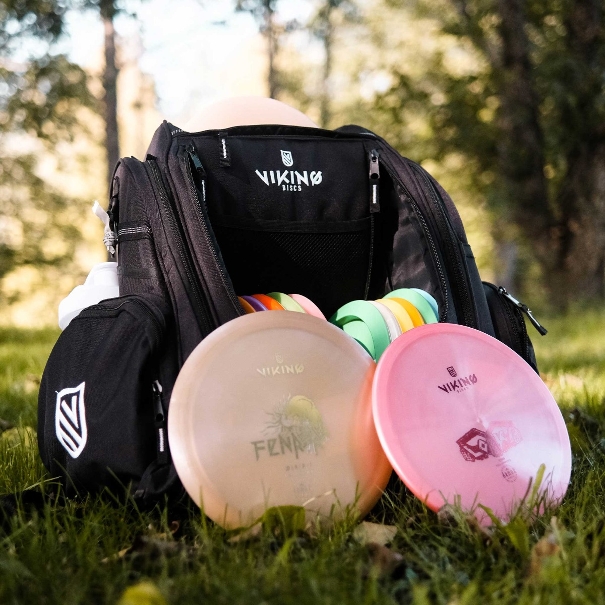 Schwarze Disc-Golf-Tasche offen mit Scheiben darin. Zwei Scheiben sind vorne, beige und rosa. Grüner Rasenhintergrund.