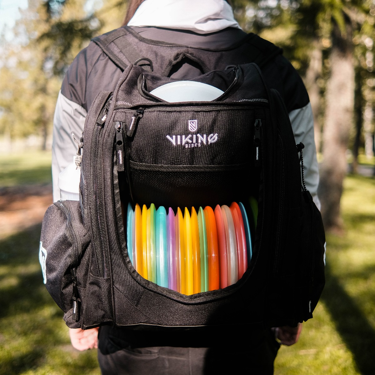 Eine Person mit einem Rucksack voller bunter Discgolf-Scheiben in einer grünen, sonnigen Umgebung.