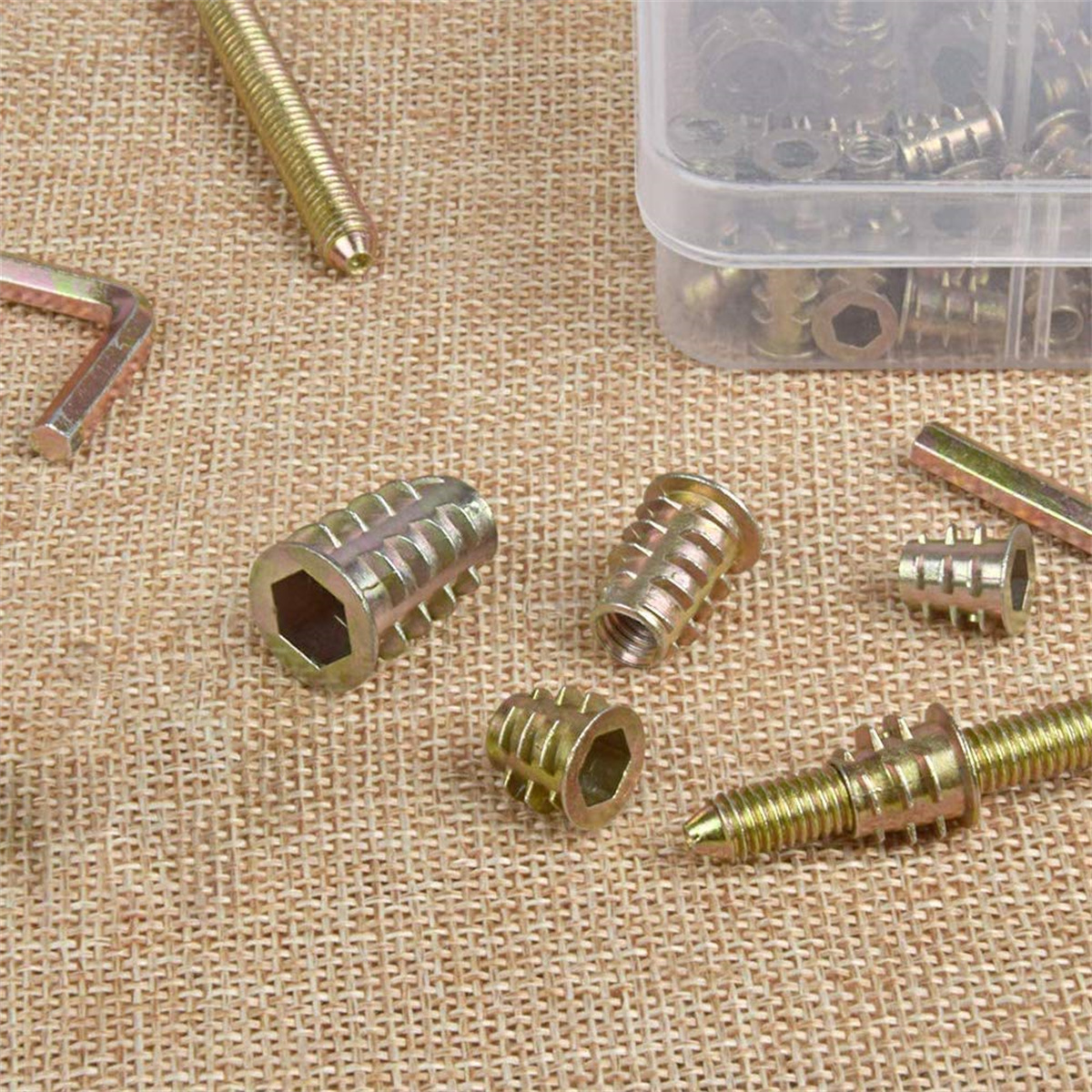 Verschiedene Metallbefestigungselemente, darunter Schrauben und Einschraubmuttern, auf einer rauen, beigefarbenen Stoffoberfläche.