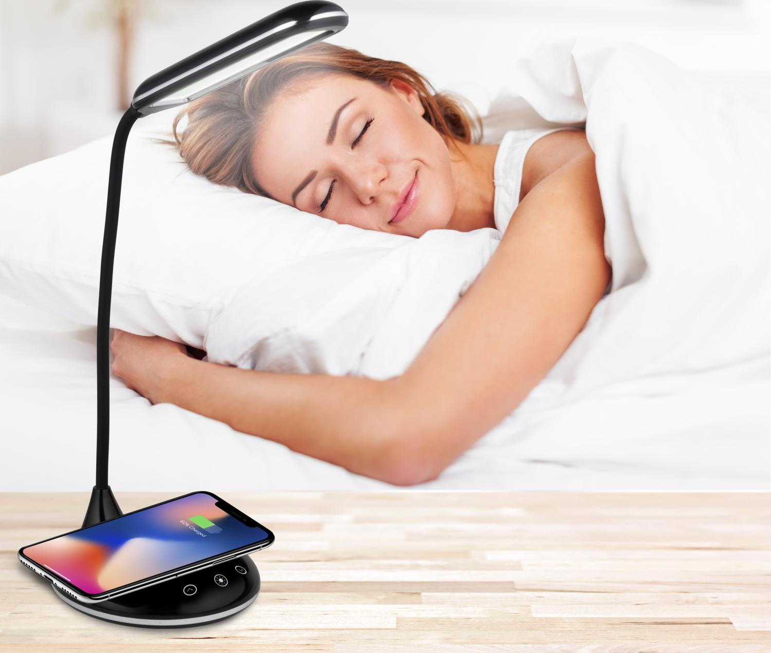 A woman sleeps in bed with a lamp over the head. A phone is on the bedside table, charging.