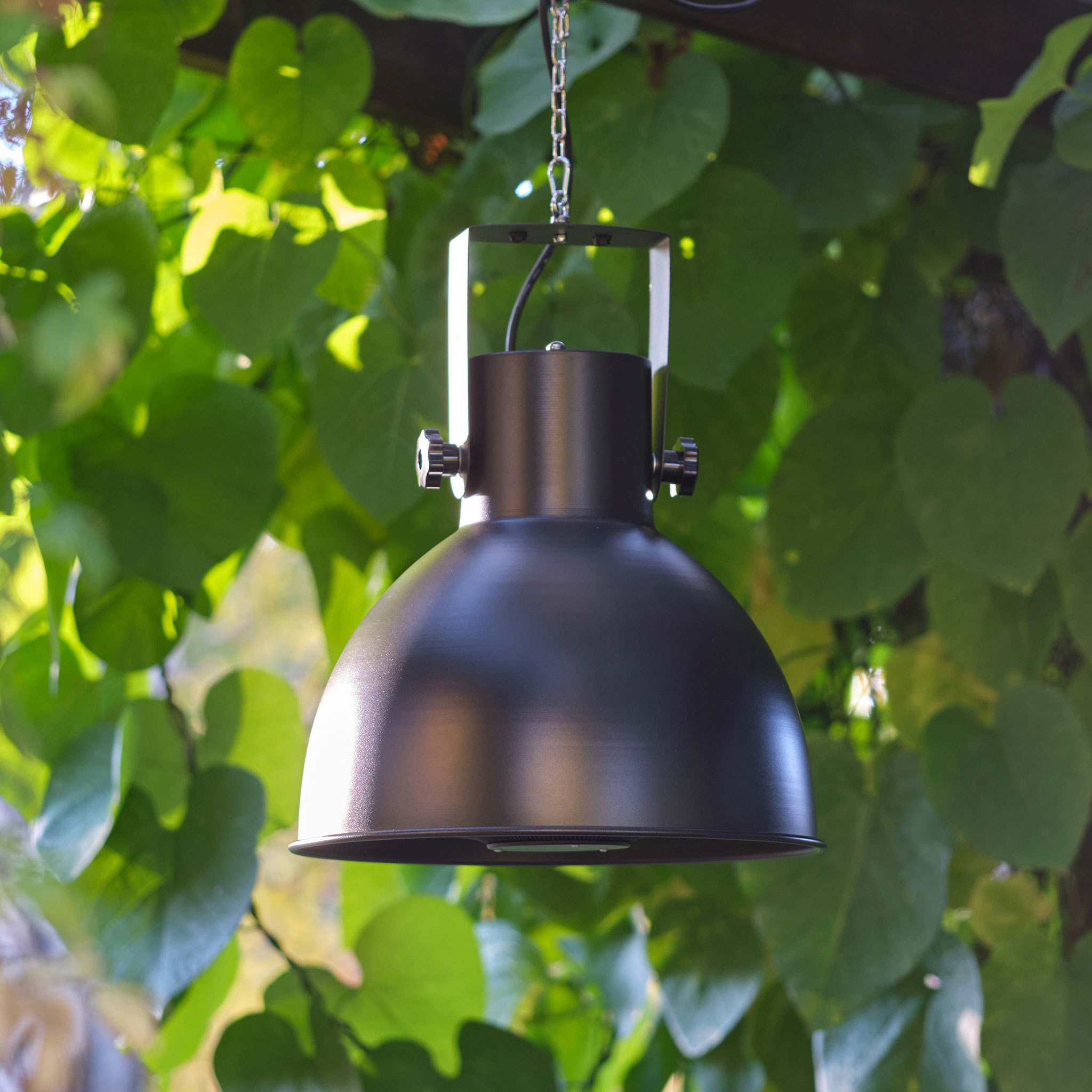 Eine schwarze Außenlampe, die an einer Kette hängt, umgeben von grünem Laub.