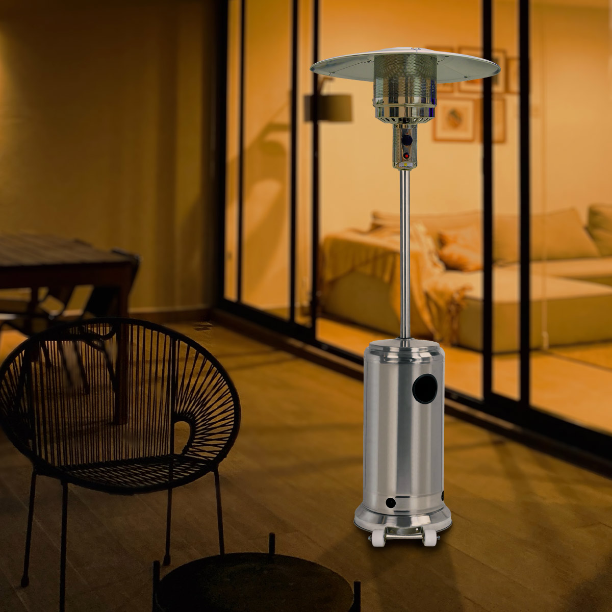 Zewnętrzny srebrny ogrzewacz tarasowy na drewnianym tarasie. Zawiera grzejnik, czarny fotel i stół, z rozmytym tłem.