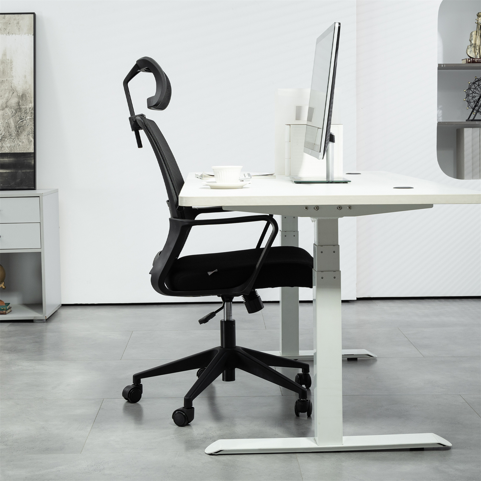Ein schwarzer Bürostuhl neben einem weißen Schreibtisch mit Monitor. Weißer Raum, natürliches Licht.