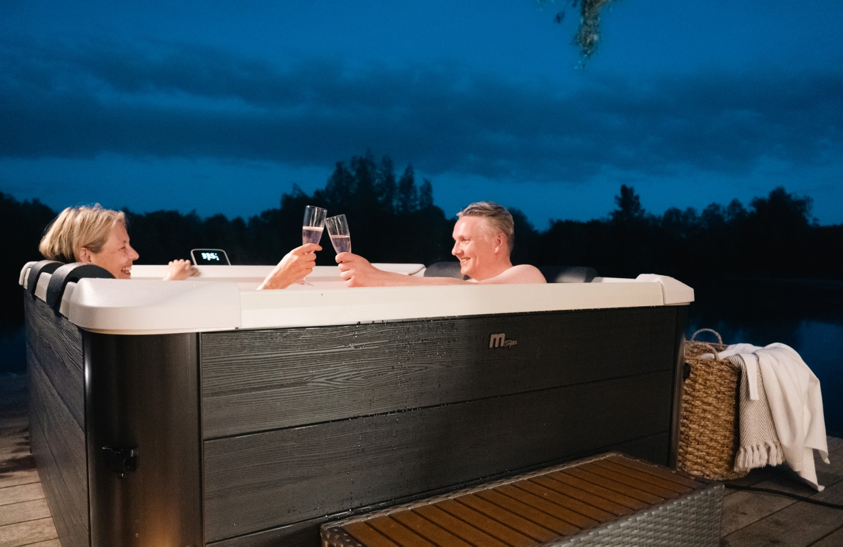 Ein Paar prostet sich mit Champagner in einem Whirlpool bei Nacht zu. Dunkle Landschaft im Hintergrund.