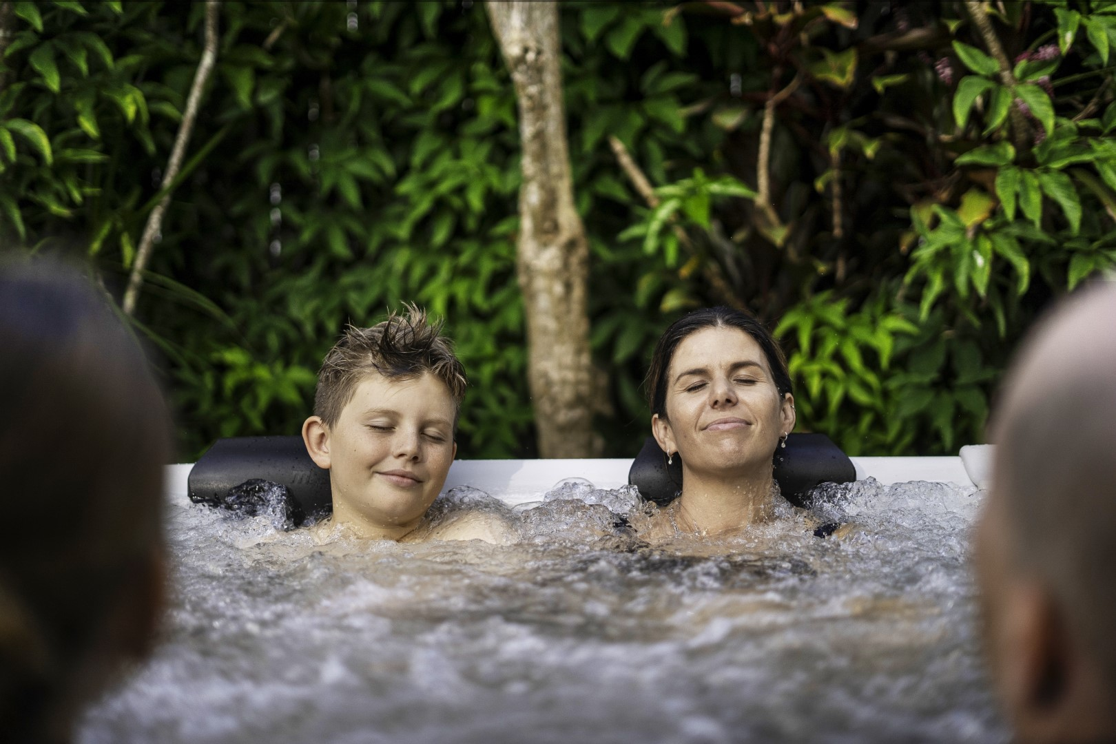 Menschen, die sich im Freien in einem Whirlpool entspannen, umgeben von üppigem Grün, die Augen geschlossen.