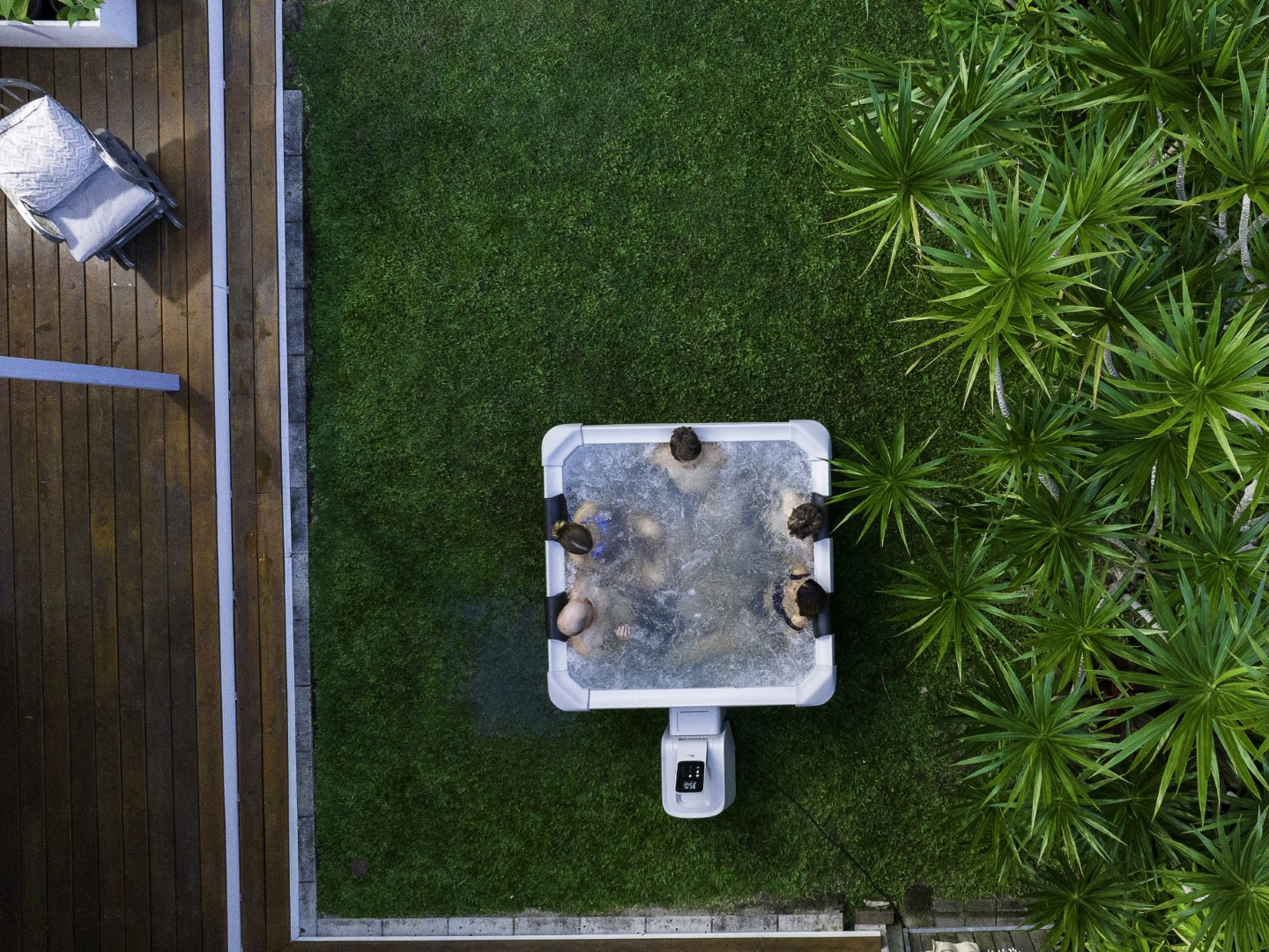 Draufsicht auf Personen in einem weißen Whirlpool auf grünem Gras. Pflanzen und Holzdeck umgeben es.