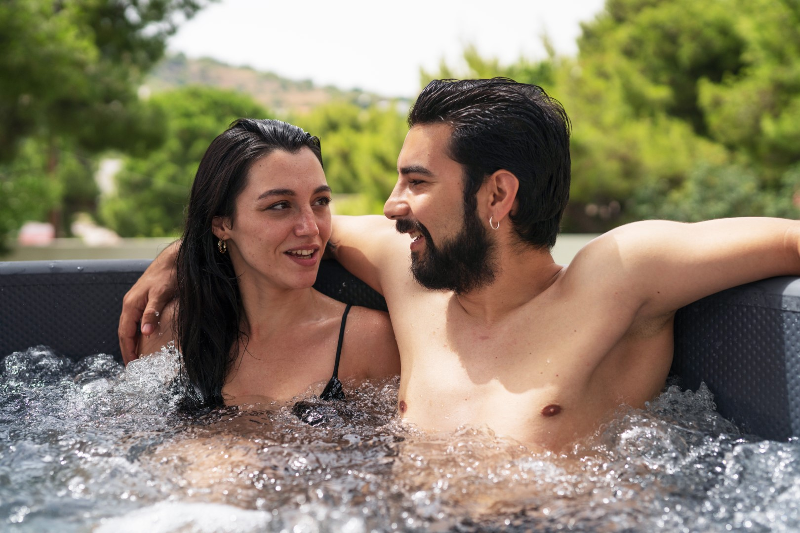 Ein Paar in einem Whirlpool mit klarem Wasser. Sie lächeln. Der Hintergrund umfasst Bäume.
