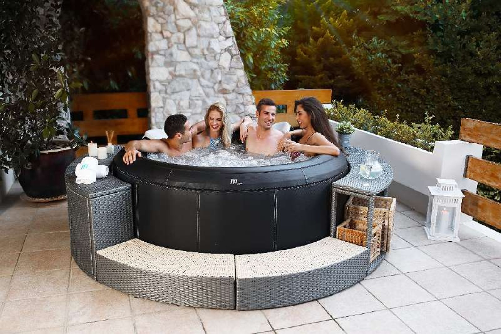Vier Personen in einem Whirlpool auf einer Terrasse, umgeben von einer schwarzen Wanne und Korbmöbeln.