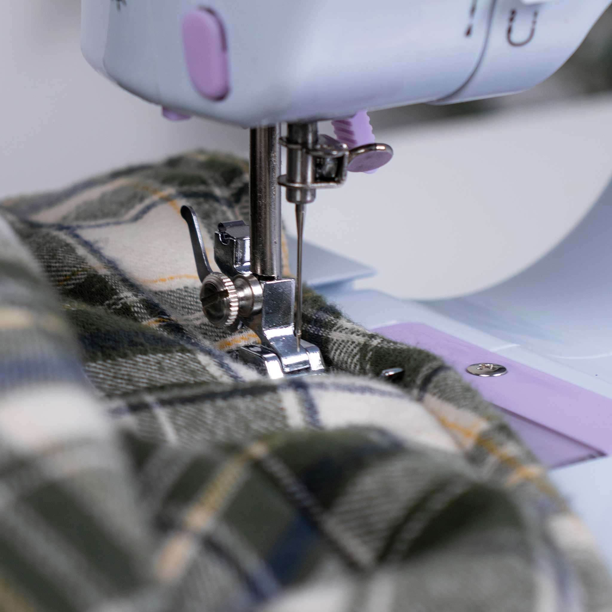 Igła maszyny do szycia przeszywająca tkaninę w kratę. Maszyna jest biała i fioletowa.