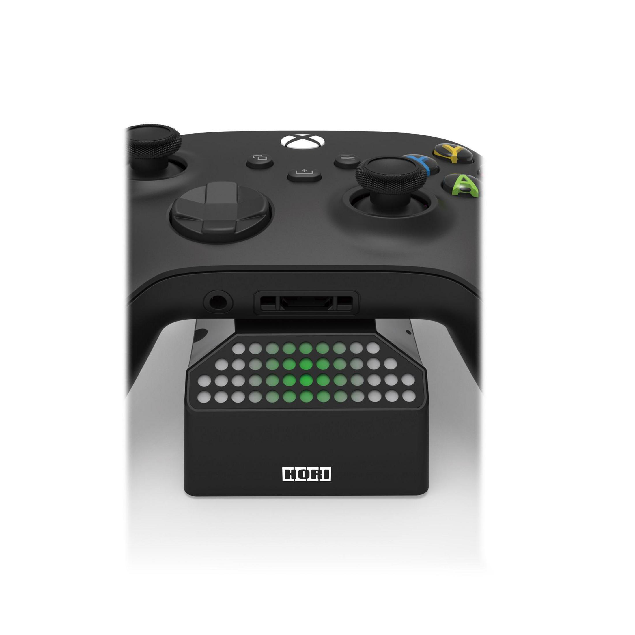 Schwarzer Xbox-Controller auf einer Ladestation. Grüne Lichter, weißer Hintergrund.