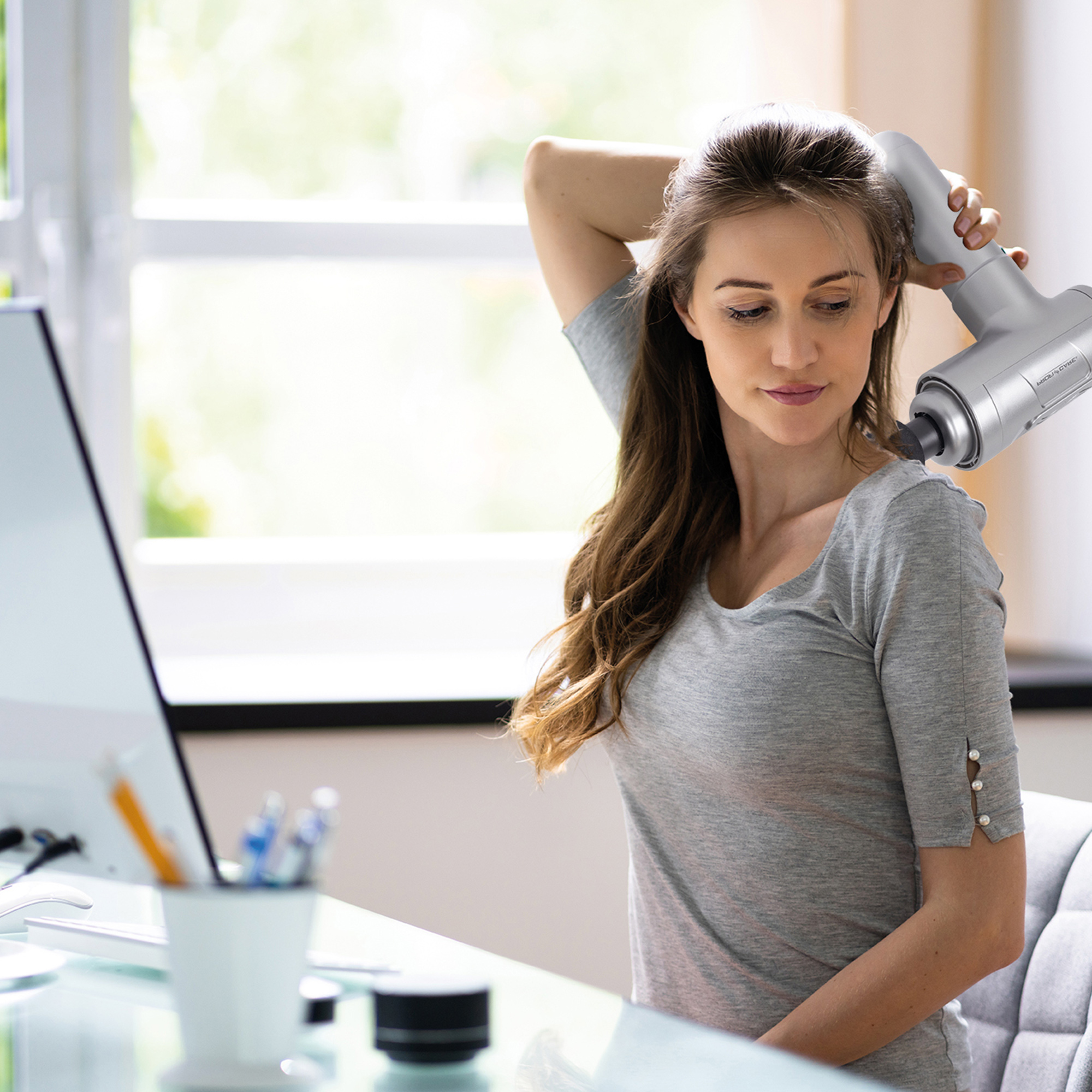 Kobieta używająca pistoletu do masażu na szyi podczas pracy przy komputerze w biurze.