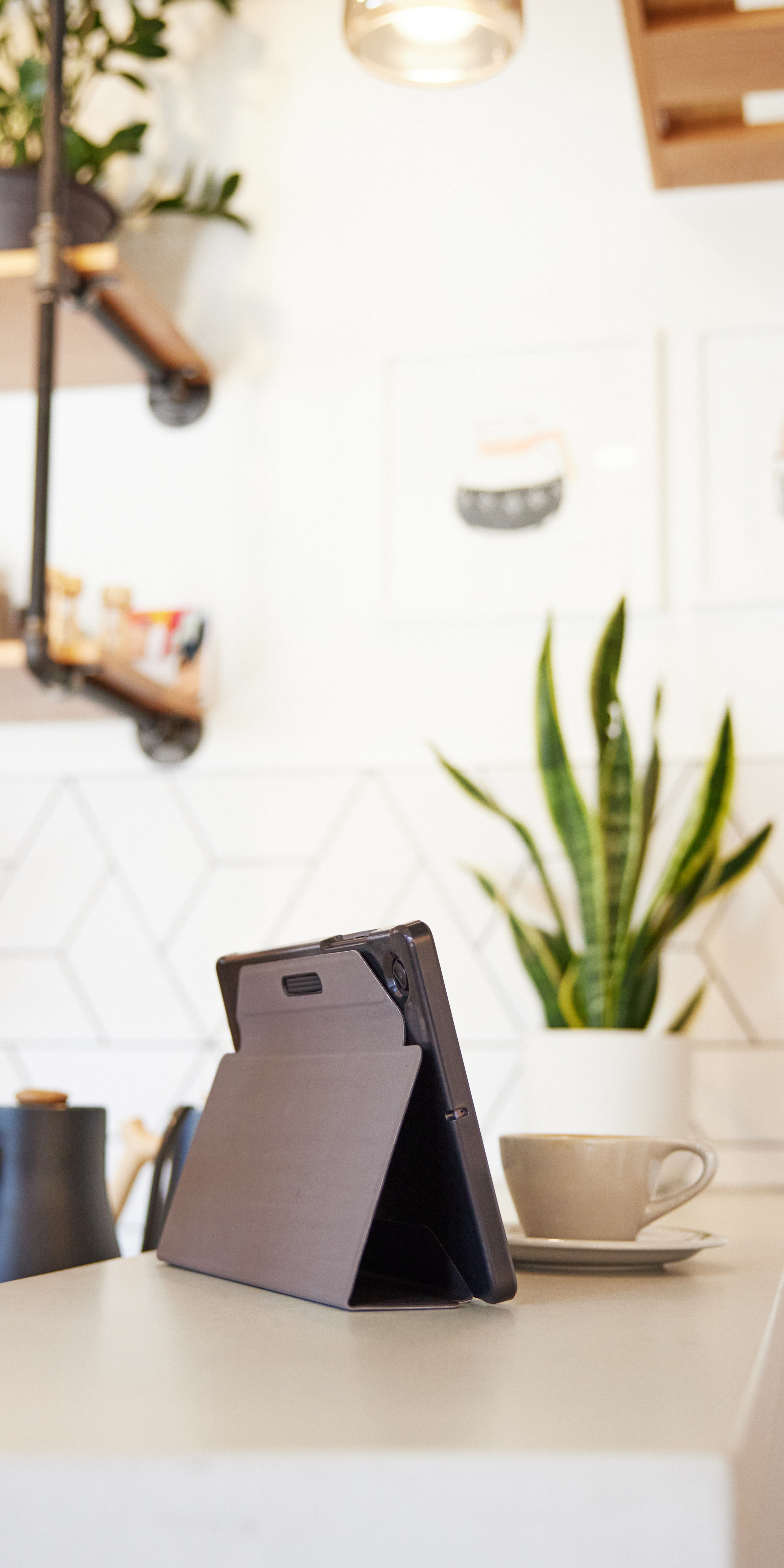 Ein Tablet in einem Gehäuse steht auf einer Theke. Eine Kaffeetasse und eine Pflanze im Hintergrund.