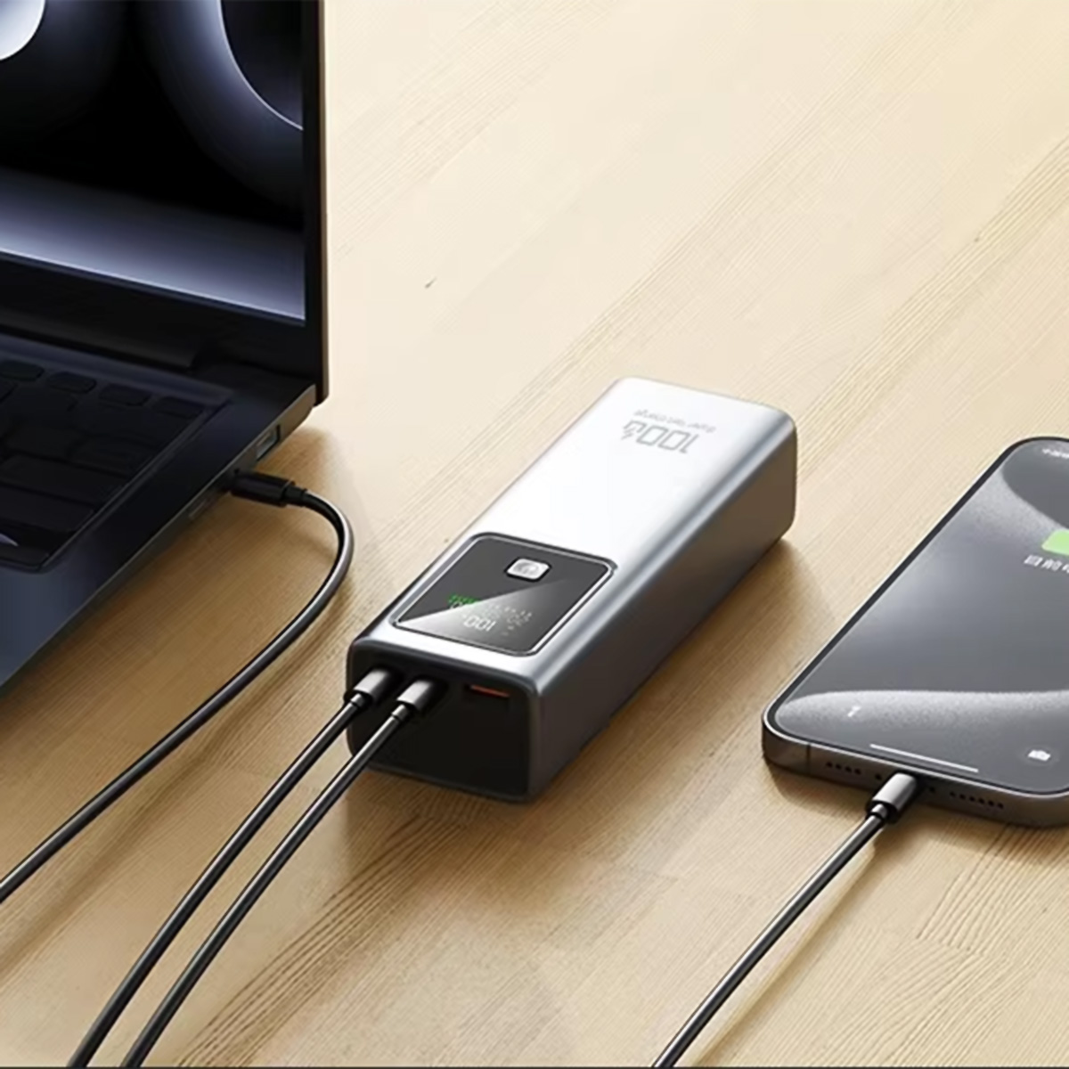 Banco de energía plateado cargando un portátil y un teléfono, sobre superficie de madera.