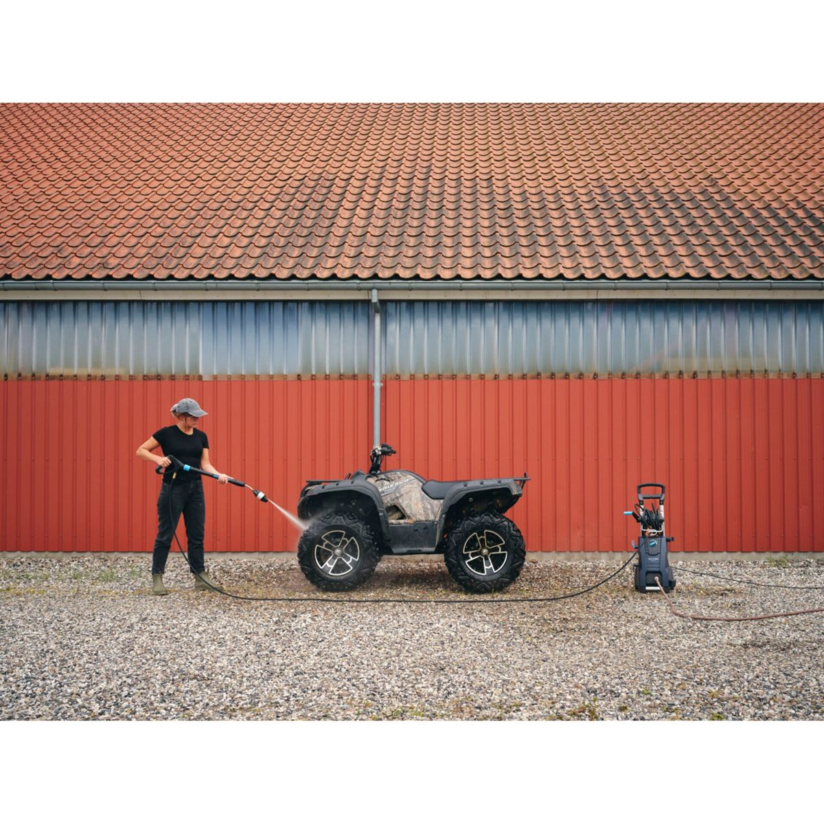 Une femme lave un VTT avec un nettoyeur haute pression. Bâtiment rouge en arrière-plan. Sol en gravier.