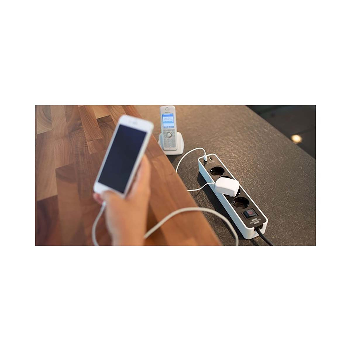 Mano che tiene un telefono con schermo nero, vicino a una presa multipla e un telefono cordless.
