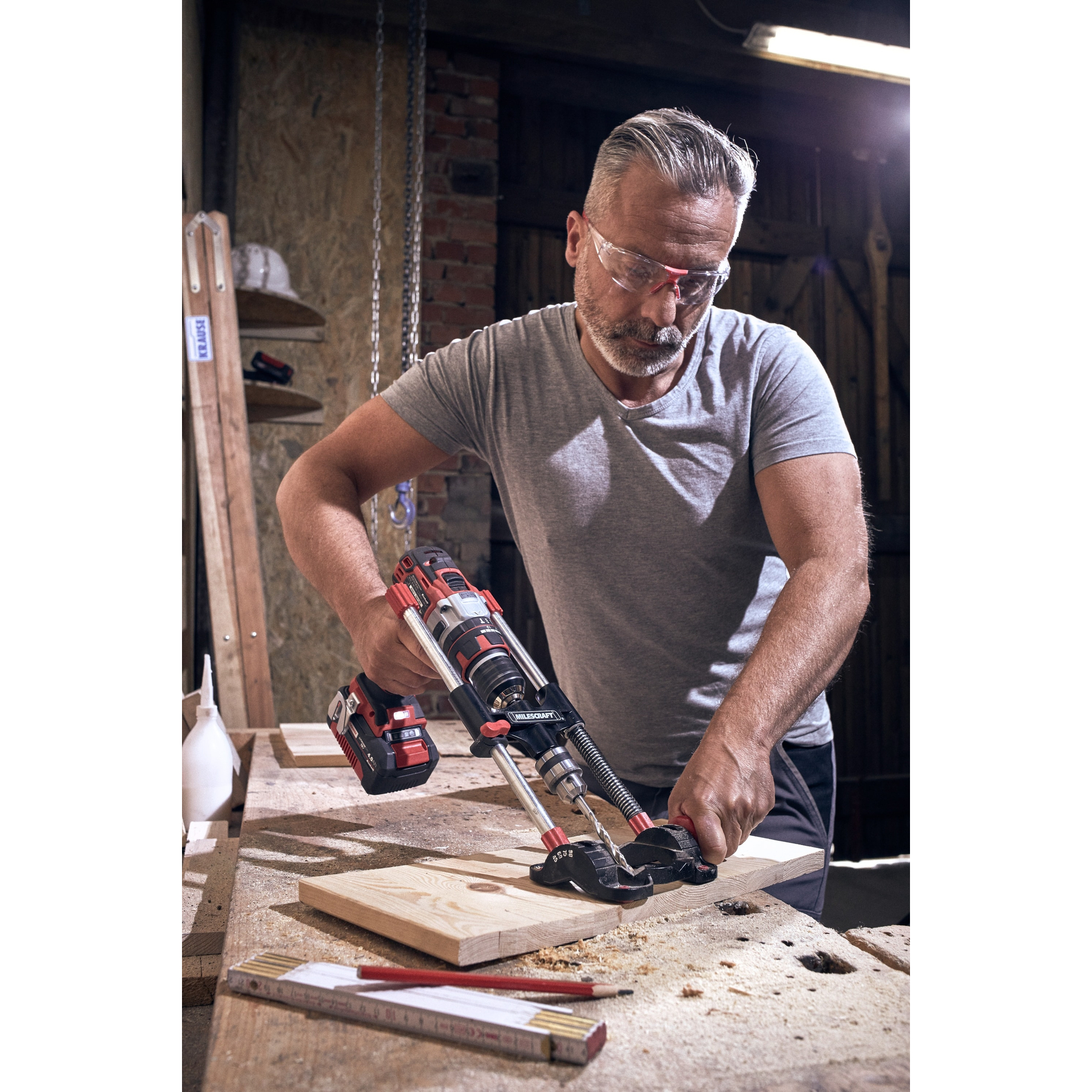 Ein Mann mit Schutzbrille benutzt eine Bohrmaschine auf einem Holzbrett in einer Werkstatt.