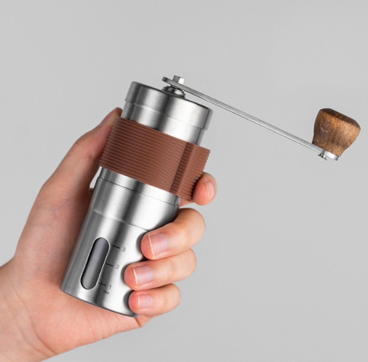Eine Hand hält eine silberne und braune manuelle Kaffeemühle auf hellgrauem Hintergrund.