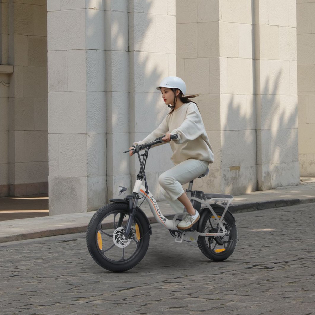 Kobieta jedzie rowerem elektrycznym na ulicy z budynkami po obu stronach.