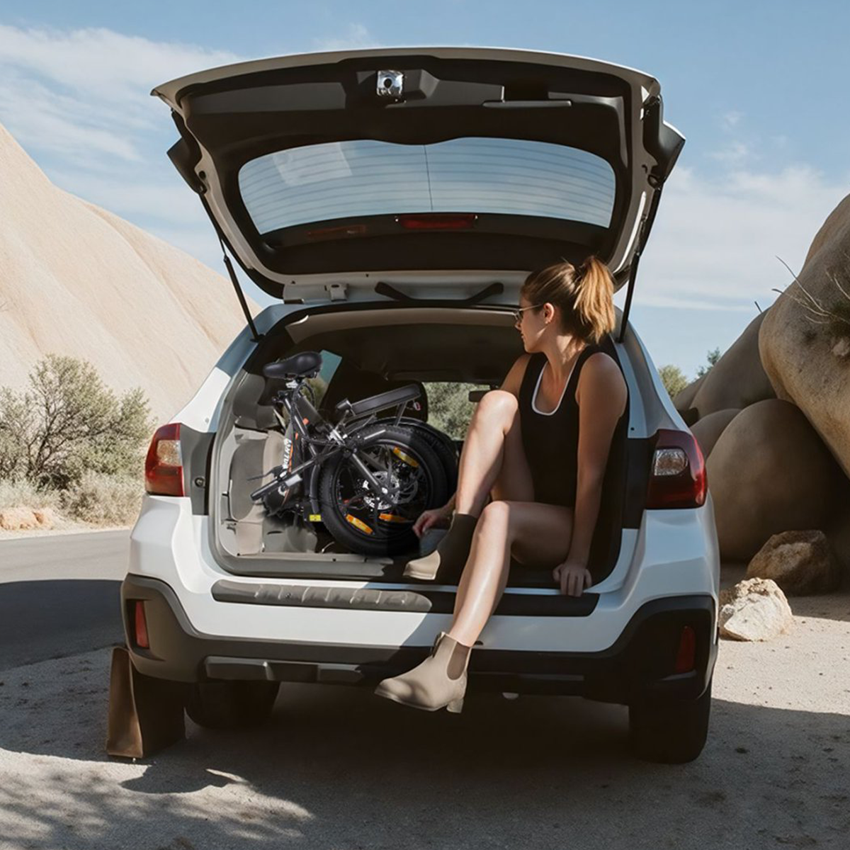 Kobieta siedząca w bagażniku samochodu ze złożonym czarnym rowerem. Tło: krajobraz pustynny.