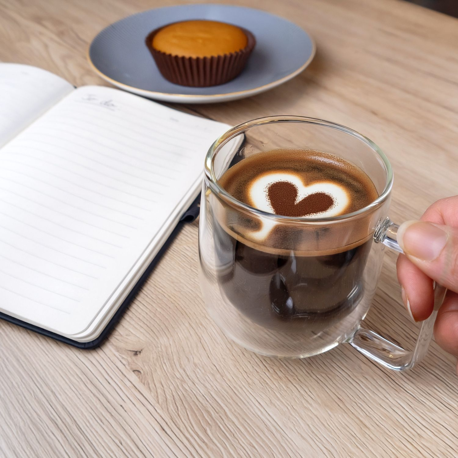 Kaffee in einem durchsichtigen Becher mit herzförmigem Design, neben einem Notizbuch und Muffin.
