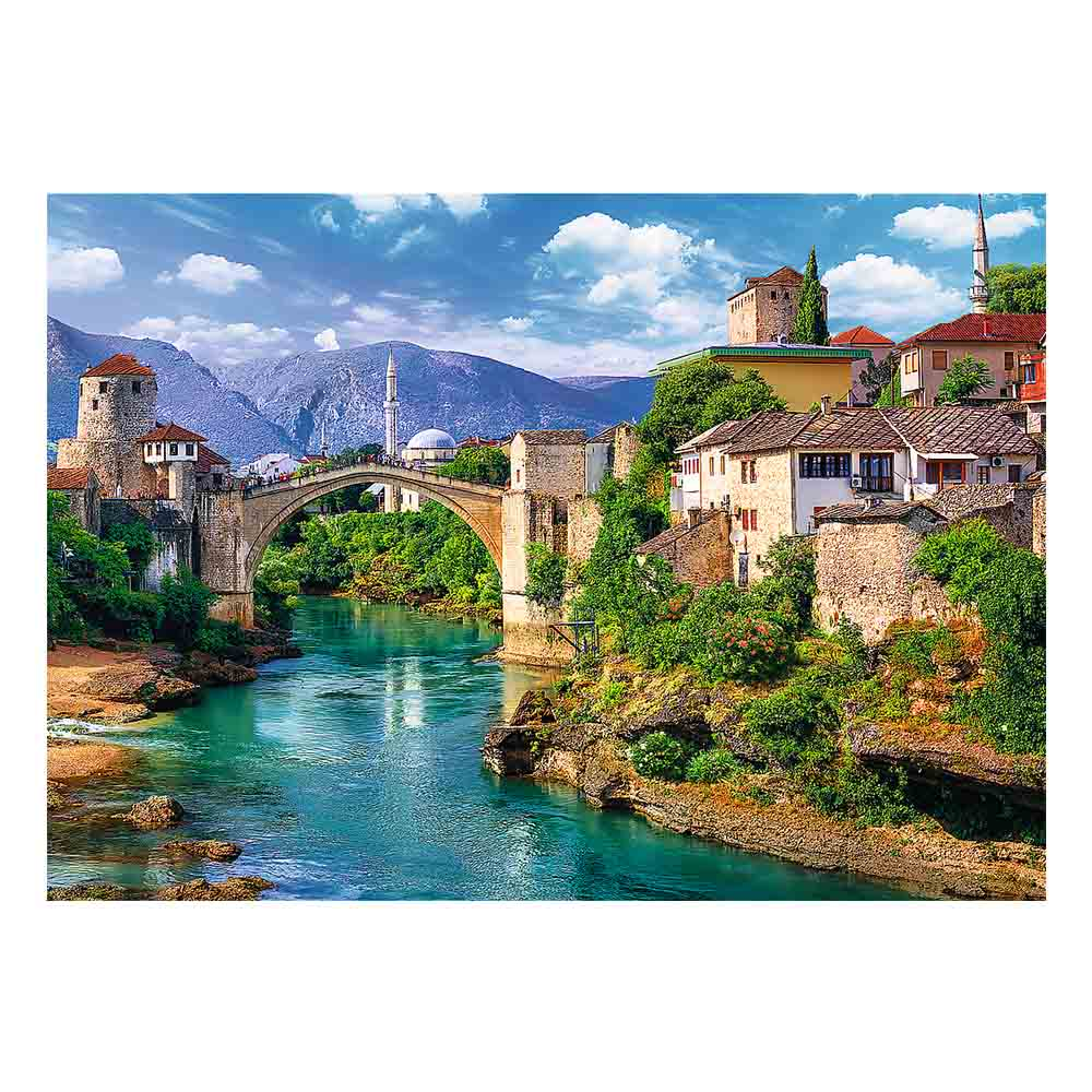 Most nad turkusową rzeką, z budynkami po obu stronach. W tle góry i błękitne niebo.