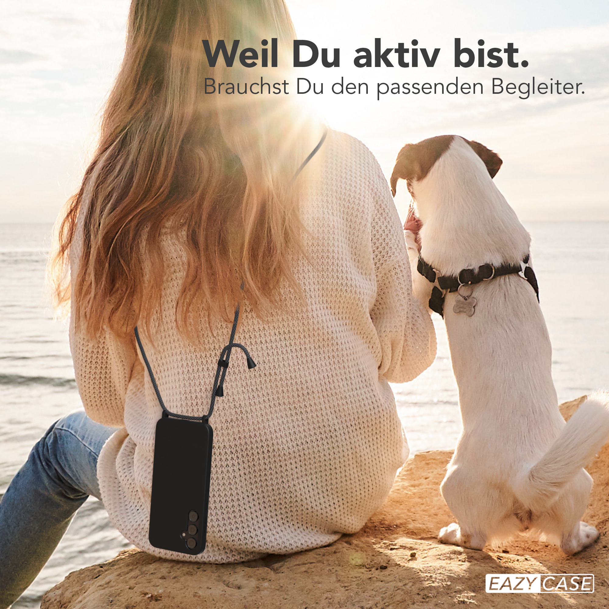 Eine Frau und ein Hund am Strand. Die Frau hält ein Telefon mit einem Umhängeband.